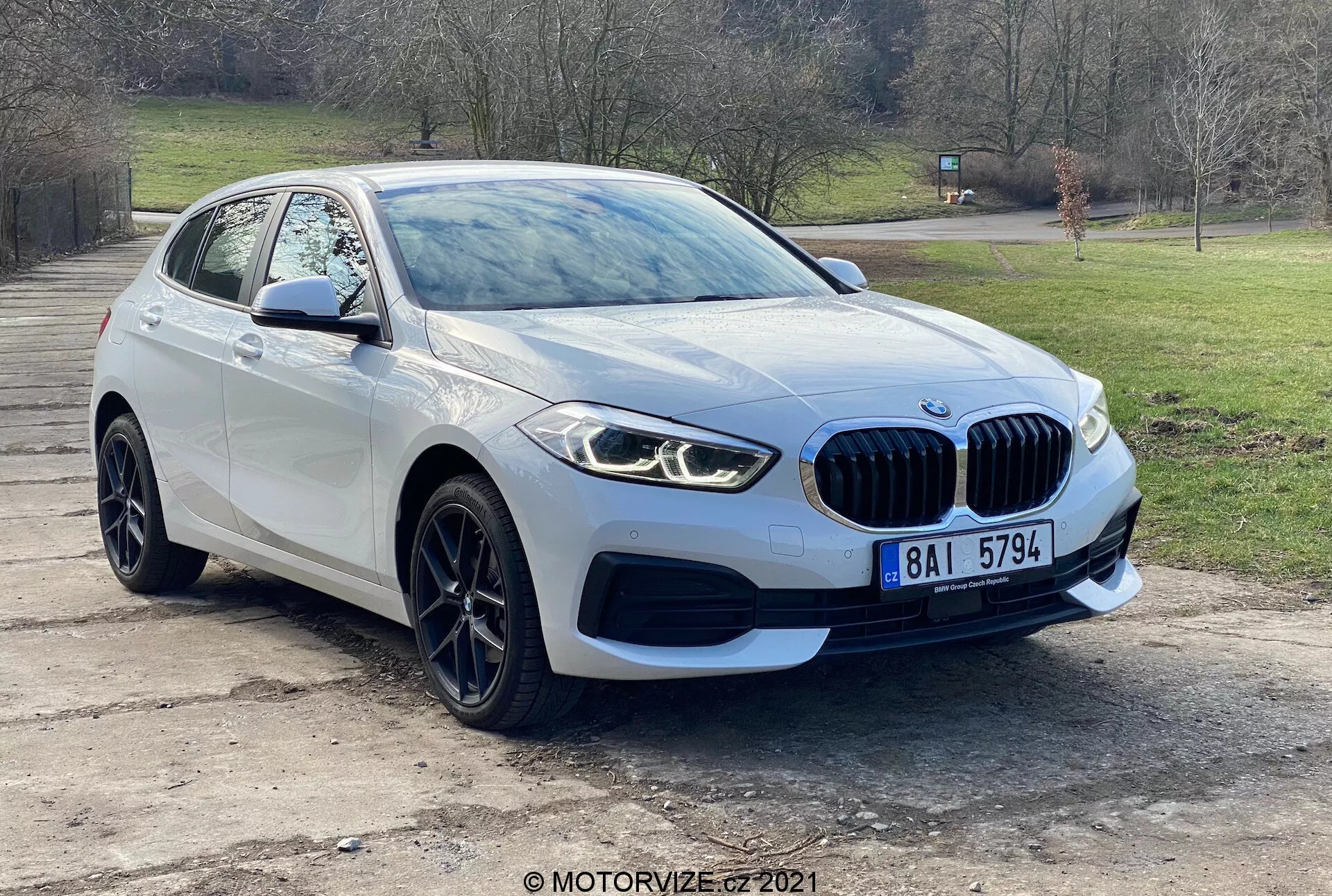 Modern BMW 116i från 2019 års 1-serie fotograferad från ett trekvartsperspektiv framifrån, som visar grill med emblem, eleganta LED-strålkastare, dimljus, främre stötfångare och aerodynamiska motorhuvslinjer som leder till vindrutan. Förarens sida av hjulet och stänkskärmen, tillsammans med sidobackspegeln med integrerade blinkers, framhäver fordonets avancerade design och parkeringssensorsteknik. Fotograferad utomhus med träd och gräs i bakgrunden, bilen har lättmetallfälgar och en distinkt njurgrill som indikerar lyx och prestanda.