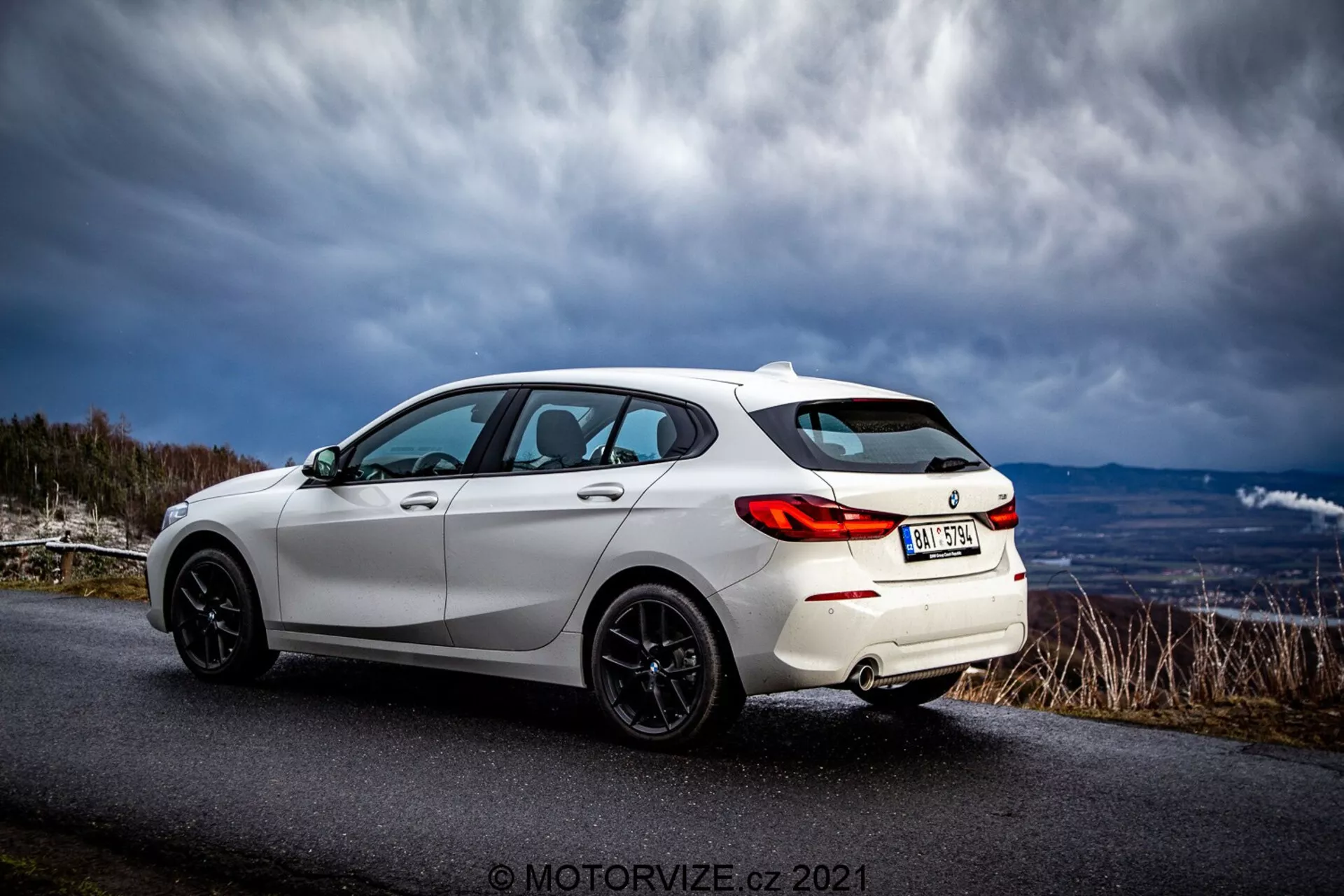 En vit, modern hatchback BMW 116i 2019 fångad från en tre-kvarts bakre vy. Bilens sportiga exteriöra egenskaper inkluderar LED-bakljus, svarta flerspakiga lättmetallfälgar, en bakspoiler, tonade fönster och eleganta karosserilinjer som framhäver bilens aerodynamiska design. Fordonet är parkerat på en asfalterad väg i ett lantligt eller bergigt område under en dramatisk himmel.