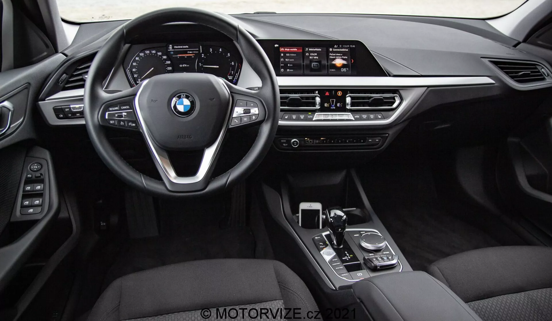 Interiörbild av en BMW 1-serie 2019 (BMW 116i) tagen från baksätet, som visar hela främre instrumentpanelen, ratt med BMW-logotyp och kontrollknappar, och mittkonsol. Synliga funktioner inkluderar den breda infotainmentskärmen, manuella klimatkontroller, luftventiler, växelspak, roterande ratt och pekplatta. Interiören har svarta läder- eller konstläderytor med metalliska och högglansiga trimelement, vilket betonar lyx och avancerad förarteknik.
