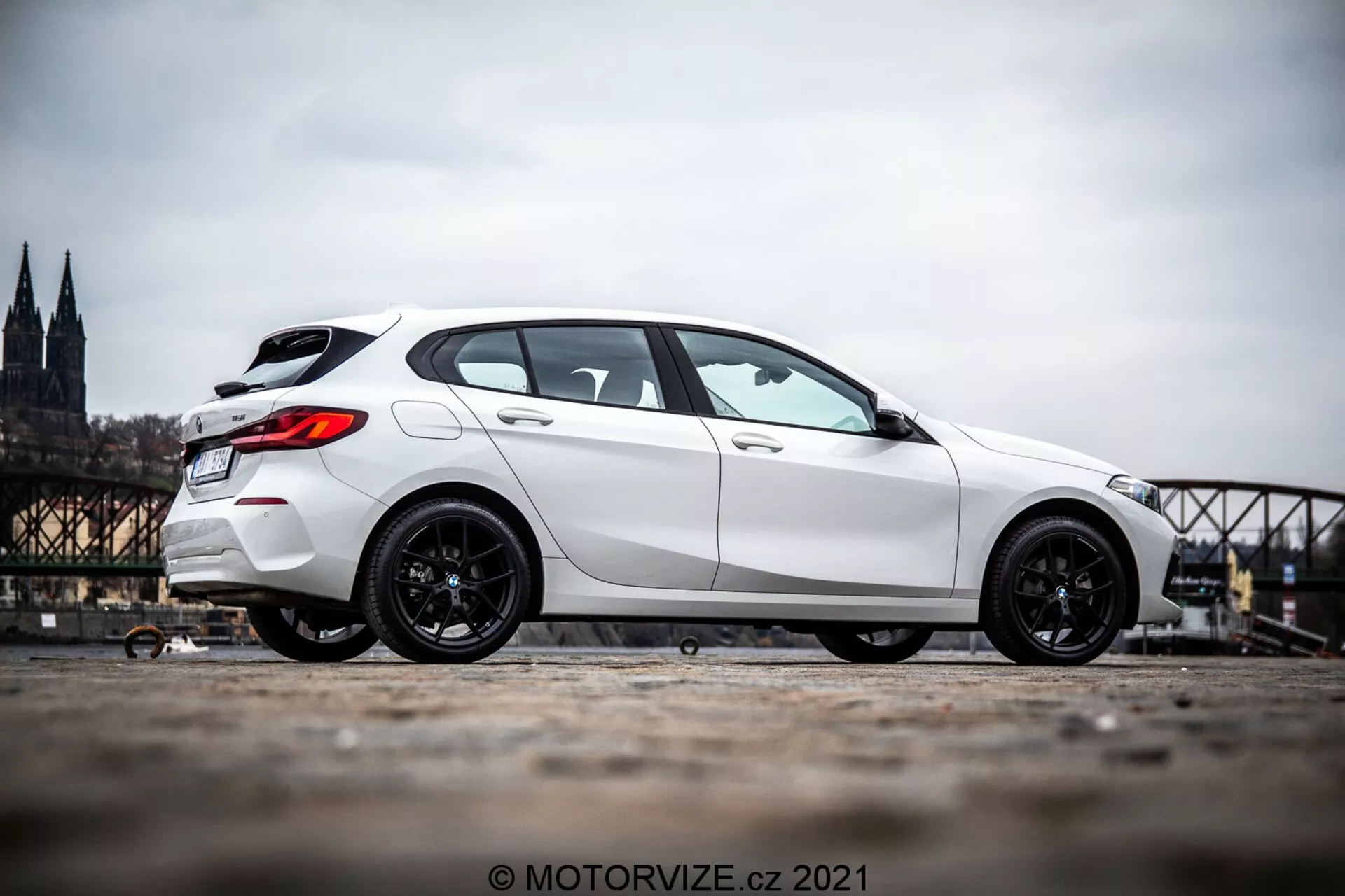 En vit 2019 BMW 1-serien (BMW 116i) halvkombi fotograferad från en profilvinkel som visar fordonets sida från strax framför framhjulet till bakre delen. Bilen är placerad på vänster sida av bilden med en suddig historisk stadsbakgrund som inkluderar en katedral och en bro. Bilden framhäver bilens sportiga mörka fälgar och dess dominerande hållning tack vare perspektivet från lägre marknivå och kamerans uppåtlutning. Fotot är taget utomhus under dagen, vilket avslöjar detaljer om bilens estetik i naturligt dagsljus.