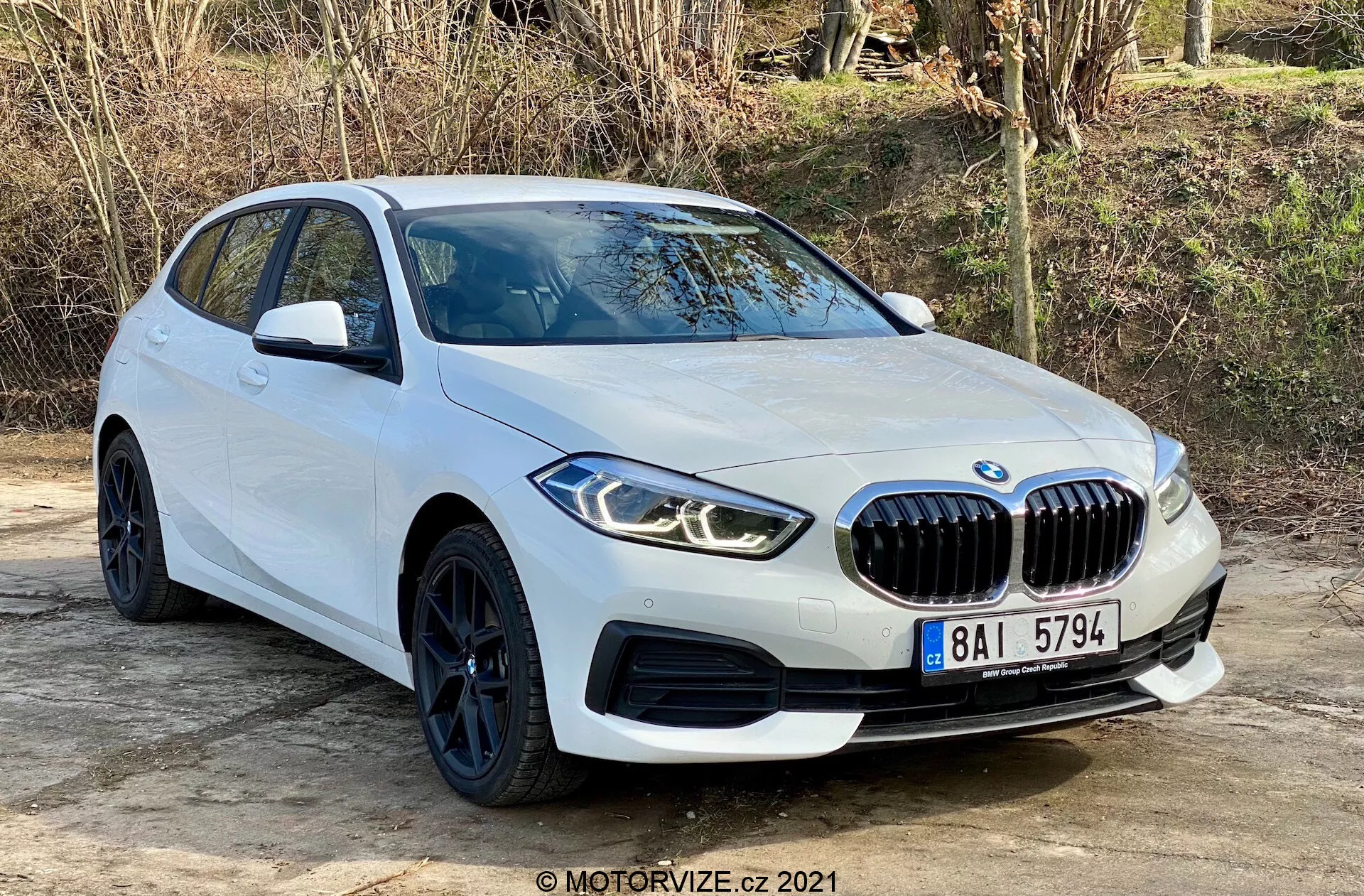 2019 BMW 116i 1-serie hatchback i vitt, fångad från en främre trekvarts vy, som visar dess framträdande njurformade frontgrill, vinklade LED-strålkastare, eleganta svarta hjul med lågprofildäck och aerodynamiska karosserilinjer. Positionerad utomhus på en plan yta med naturlig vegetation i bakgrunden, betonar bilden bilens moderna design och sportiga egenskaper, inklusive parkeringssensorer och sidospeglar med integrerade blinkers.