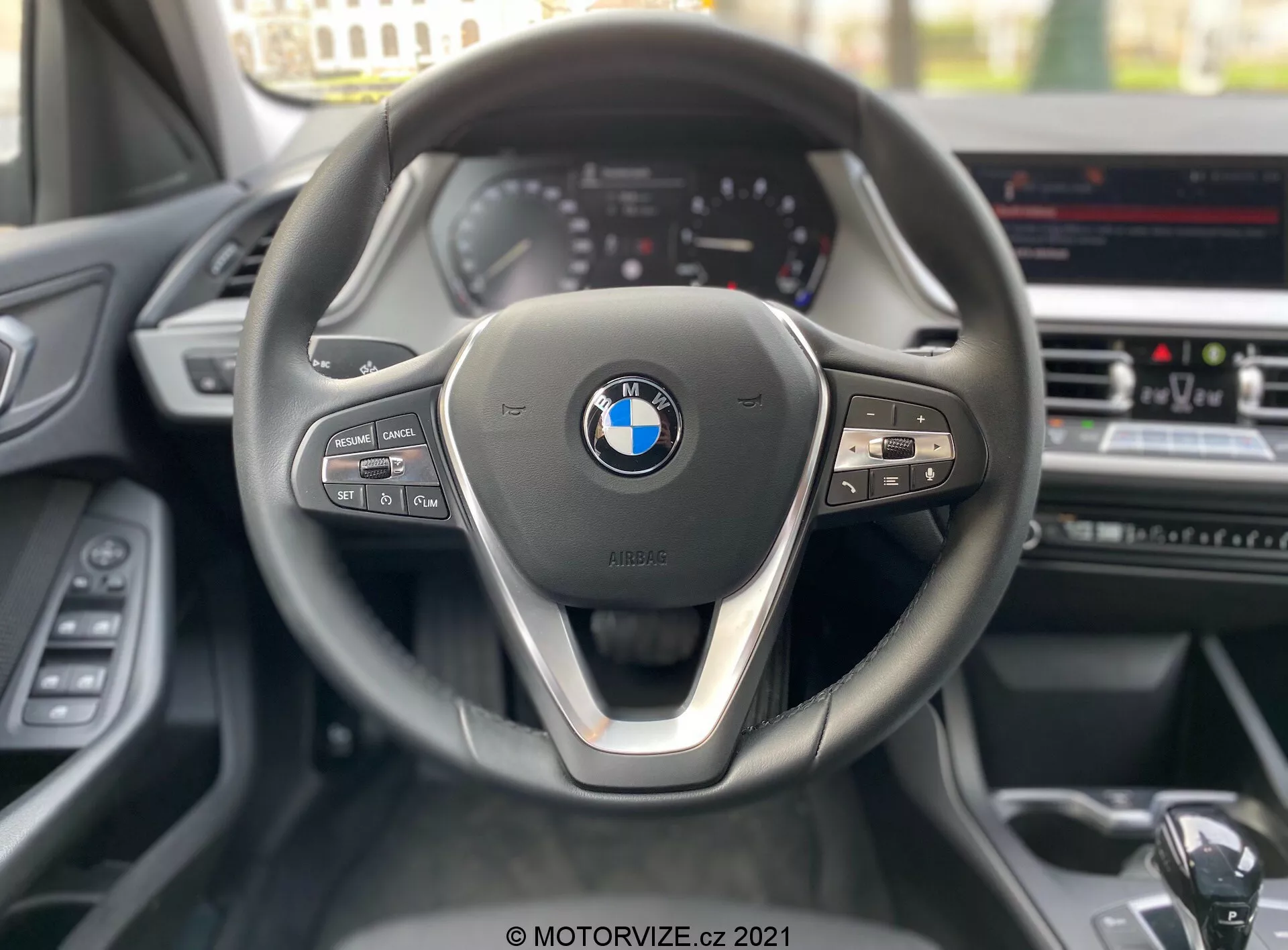 Förarperspektiv av en BMW 1-serie 2019 (BMW-116i), fokuserande på den samtida treekrade ratten med en metallaccent på den nedre ekern och den framträdande BMW-emblemet i mitten. På vänster sida av ratten finns medie- och kommunikationskontroller, medan höger sida innehåller farthållarknappar och en synlig växelspak. I bakgrunden syns en delvis synlig instrumentpanel med analoga visare och en infotainmentskärm på mittkonsolen, som antyder avancerade funktioner som GPS-navigering och klimatkontroll.