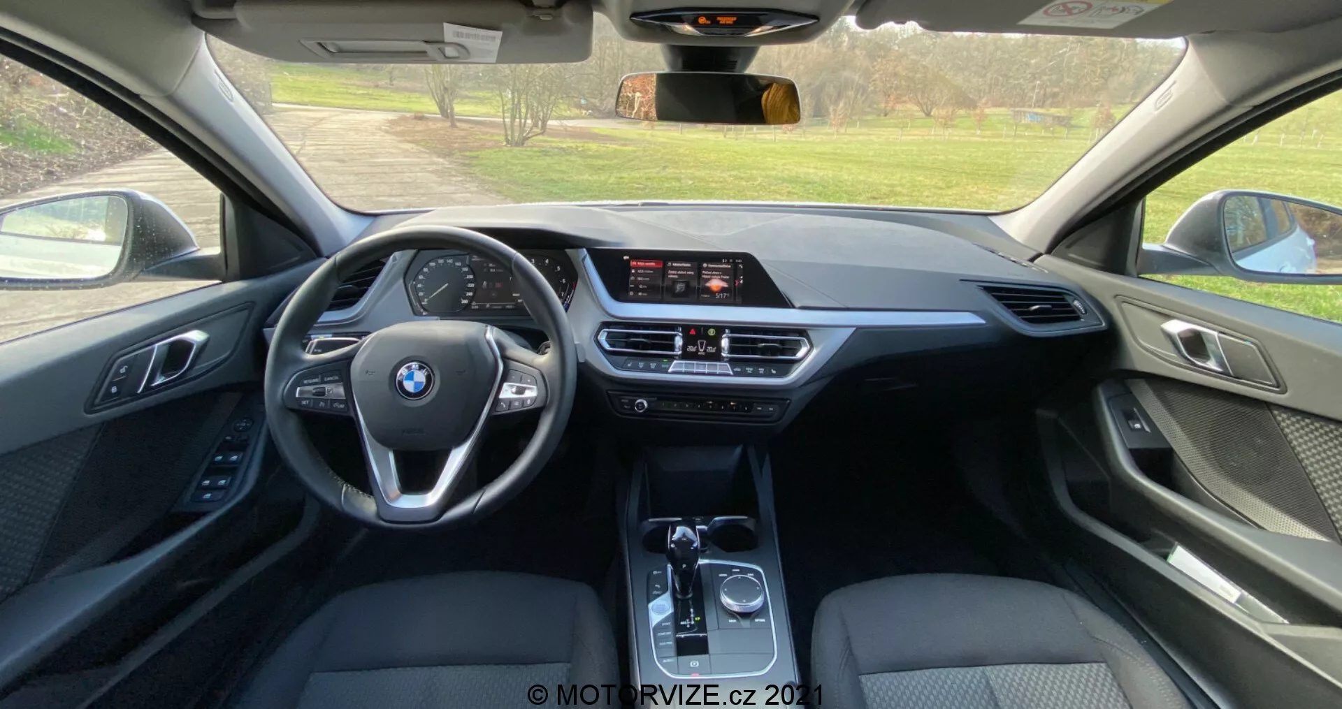 Interiörvy från baksätet av en BMW 1-serie 2019 (BMW-116i), som visar en modern hytt med en treekrad multifunktionsratt med BMW-logotypen, en fristående infotainmentskärm, tvåzons klimatanläggning och automatväxel. Användningen av läder, mjuka plaster och metalliska accenter indikerar en högnivå utrustningsgrad.