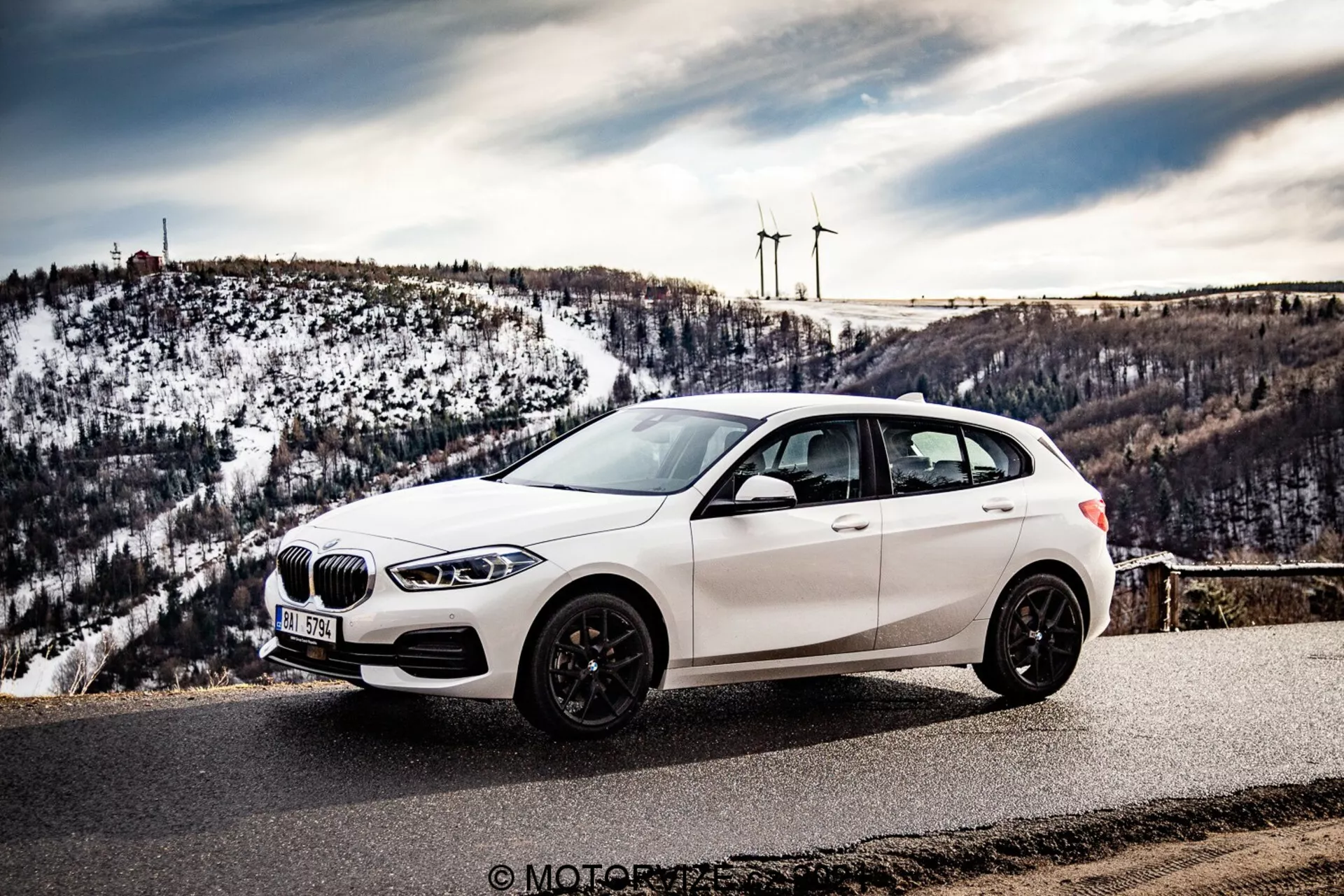 Bilden visar en vit BMW 116i hatchback från 2019 parkerad vid en naturskön utsiktsplats eller bergsväg, fotograferad från en vinkel framifrån-sidan som visar frontgrillen, LED-strålkastarna, framhjulet, sidoprofilen och en glimt av bakdelen. Perspektivet är något förhöjt, vilket framhäver bilens tak som är förskjutet till vänster, med bakgrund av snötäckta kullar och avlägsna vindkraftverk. Fordonet har ett njurformat galler, lågprofil-däck på flerspårslegeringshjul och mörkfärgade fälgar, sidospeglar med integrerade blinkers och tonade rutor. Miljön antyder klart väder, vilket förbättrar den marknadsföringsmässiga och visuellt tilltalande karaktären av fotot, troligen taget under en bilresa eller en fordonsrecension.