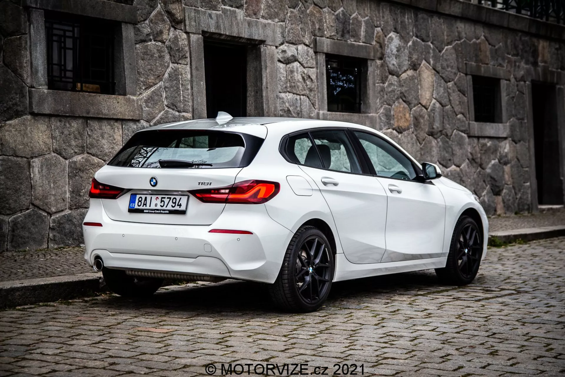 Medeldistansbild på marknivå av en vit BMW 1er-serie 116i hatchback från 2019, tagen från en tre-kvarts bakre vinkel, framträdande med bakljusens LED-design, bakrutan och sportiga fälgar. Synliga funktioner inkluderar sensorer på bakre stötfångaren, troligen för parkeringsassistans, en liten takantenn sannolikt för GPS eller satellitradio, modellmärke på bakluckan, dubbelt avgassystem som antyder prestanda, och tonade fönster för integritet eller komfort.