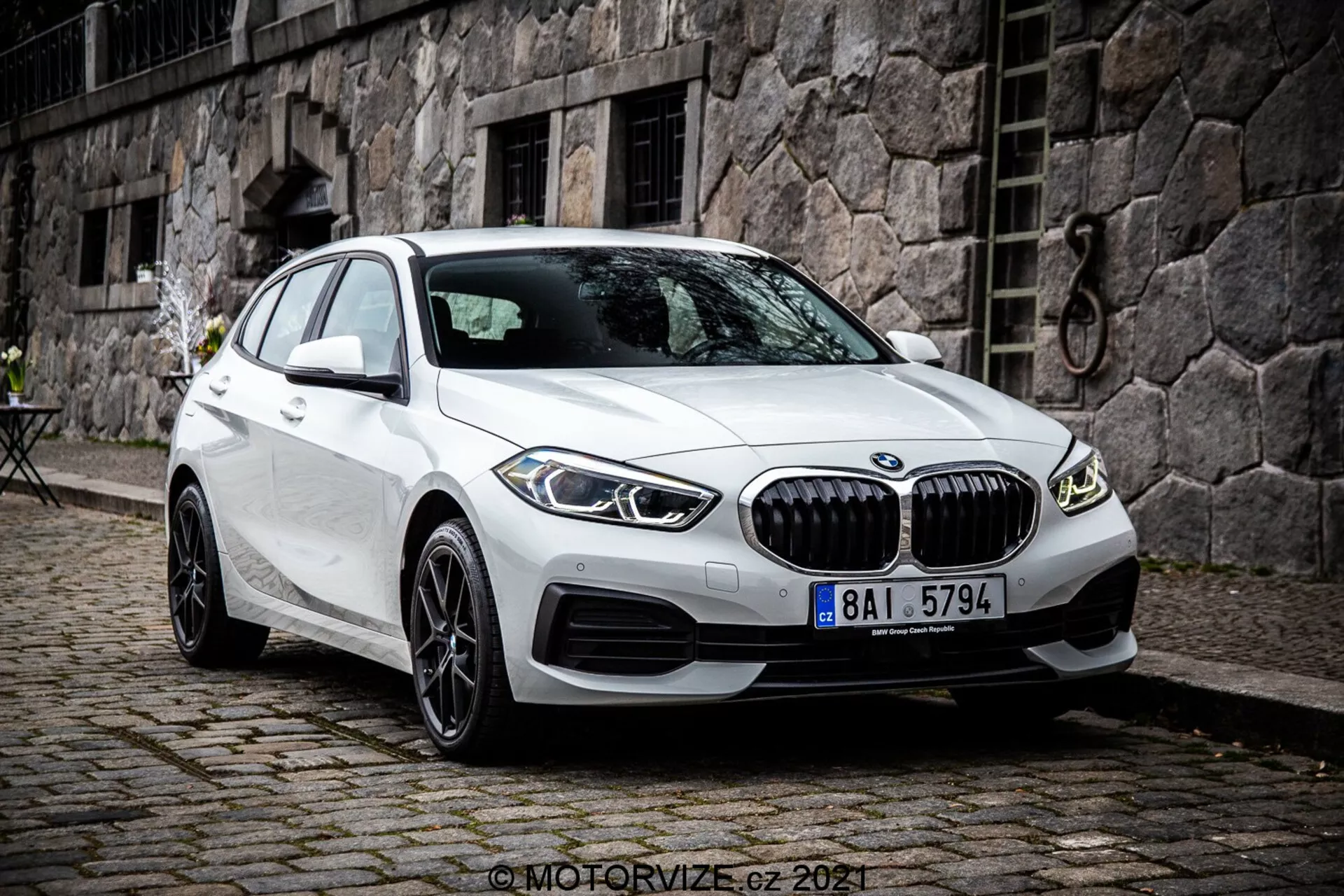 Bilden visar en BMW 1-serie från 2019, specifikt BMW 116i, i vit färg. Fotograferad från en vinkel på tre fjärdedelar framifrån, visar den bilens främre fascia, passagerarsidan och den detaljerade designen av dess hjul. Bilden tas från en något förhöjd vinkel, vilket antyder att fotografen stod eller använde ett stativ. Bilen är parkerad med framhjulen vridna åt höger, vilket ger en klar vy av fälgen och sidoprofilen på däcket. Funktionerna inkluderar en framträdande framgrill med vertikala lameller, skulpterade linjer på huven och sidopanelerna, smala LED-strålkastare och luftintag på stötfångaren som kan rymma parkeringssensorer. Sidobackspeglar med integrerade blinkers och ramlösa fönster betonar dess premium-estetik. Flerspakslegeringshjul antyder en högre utrustningsnivå eller ett valfritt tillval. Den branta bakrutan betonar hatchback-profilen. Fordonet är parkerat på kullerstenar i en urban eller historisk miljö med en stenbyggnad och ett litet bistrobord i bakgrunden. Bilden är skarp och detaljerad, tagen i naturligt mulet dagsljus.