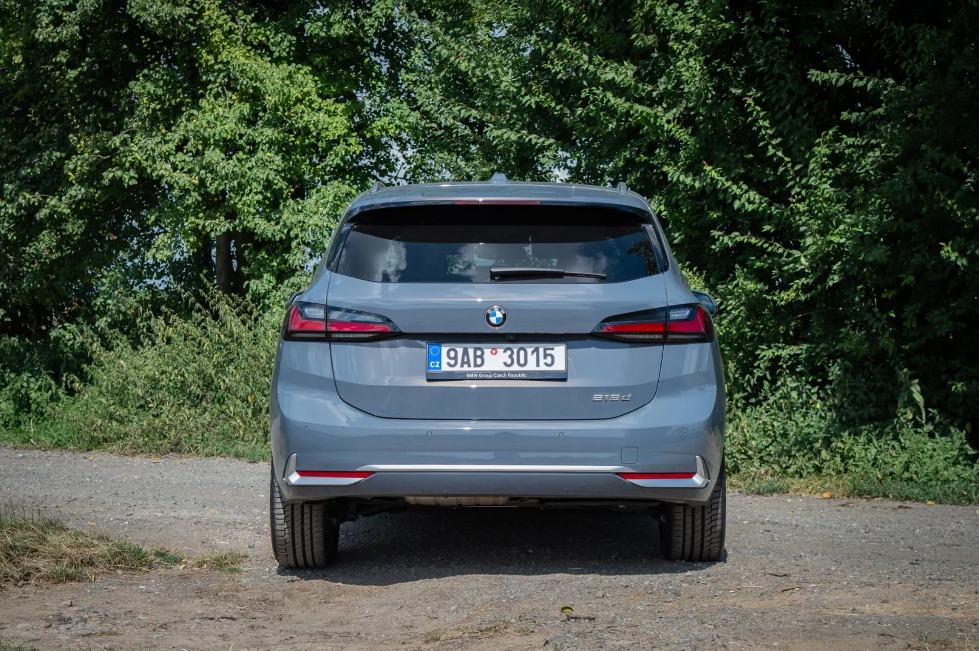 Baksida av BMW 2-serien Active Tourer 2022, som framhäver dubbla avgastoppar, parkeringssensorer bak och LED-bakljus under naturligt ljus med bakgrund av förortens grönska.