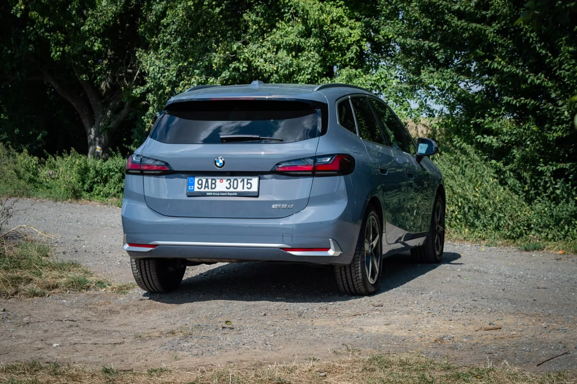 Hatchback BMW 2-serien Active Tourer 2022, sedd från en bakre vinkel något förskjuten till höger på en landsväg, med fokus på dess bakre design, LED-bakljus, dubbla avgasrör och parkeringshjälpte teknologi mot en naturlig bakgrund