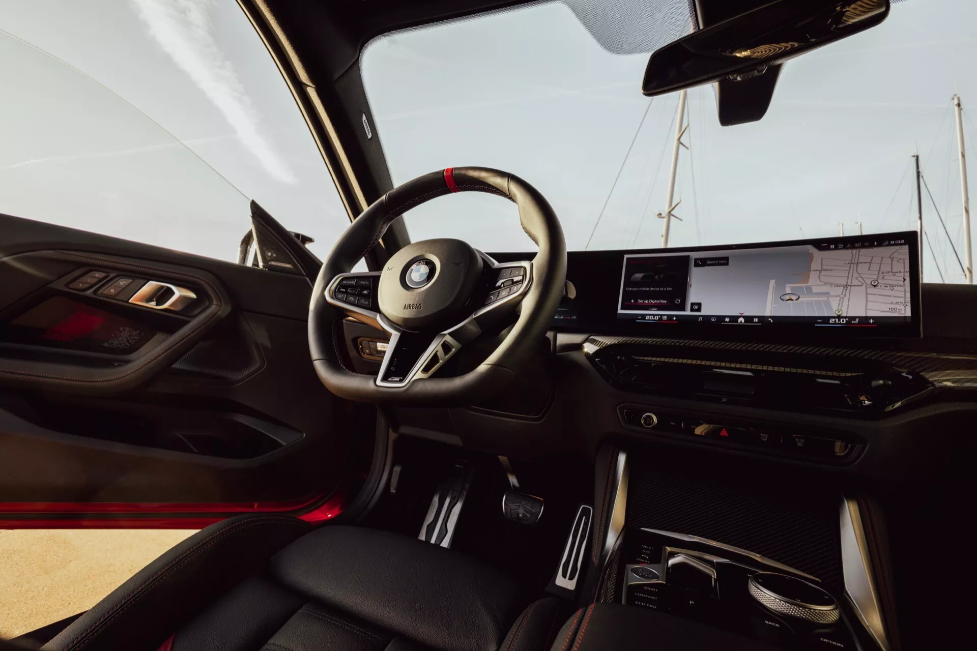 Interiör av BMW 2er-reihe 2024 (G42-G87 Facelift) från passagerarsätet. Bilden visar en treekrad ratt med integrerade kontroller, en digital instrumentpanel, en stor infotainmentskärm och minimalistiska klimatkontroller. Röd ambient belysning är synlig och växelväljare för automatisk transmission finns i mittkonsolen. Dörrpanelen har fönster- och spegelkontroller samt ett högtalargaller. Läderklädda stolar med kontrastsömmar ger en lyxig känsla.