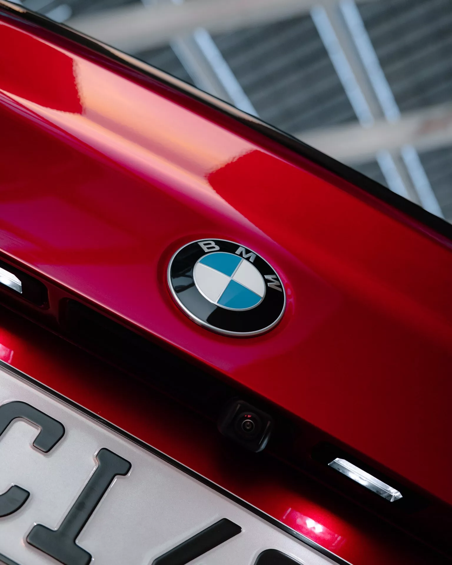 Närbild av den bakre delen av en glänsande röd 2024 BMW 2-serie (G42-G87 facelift), som visar BMW:s emblem centralt placerat på bagageluckan. Under emblemet syns en delvis synlig registreringsskylt och en liten bakåtriktad kamera som framhäver tekniska funktioner. Två små röda lampor under emblemet kan vara extra bromsljus eller bakre dimljus. Fotots uppåtvinkel och smala inramning betonar varumärkesidentiteten och de eleganta designelementen, med en bokeh-effekt i bakgrunden.