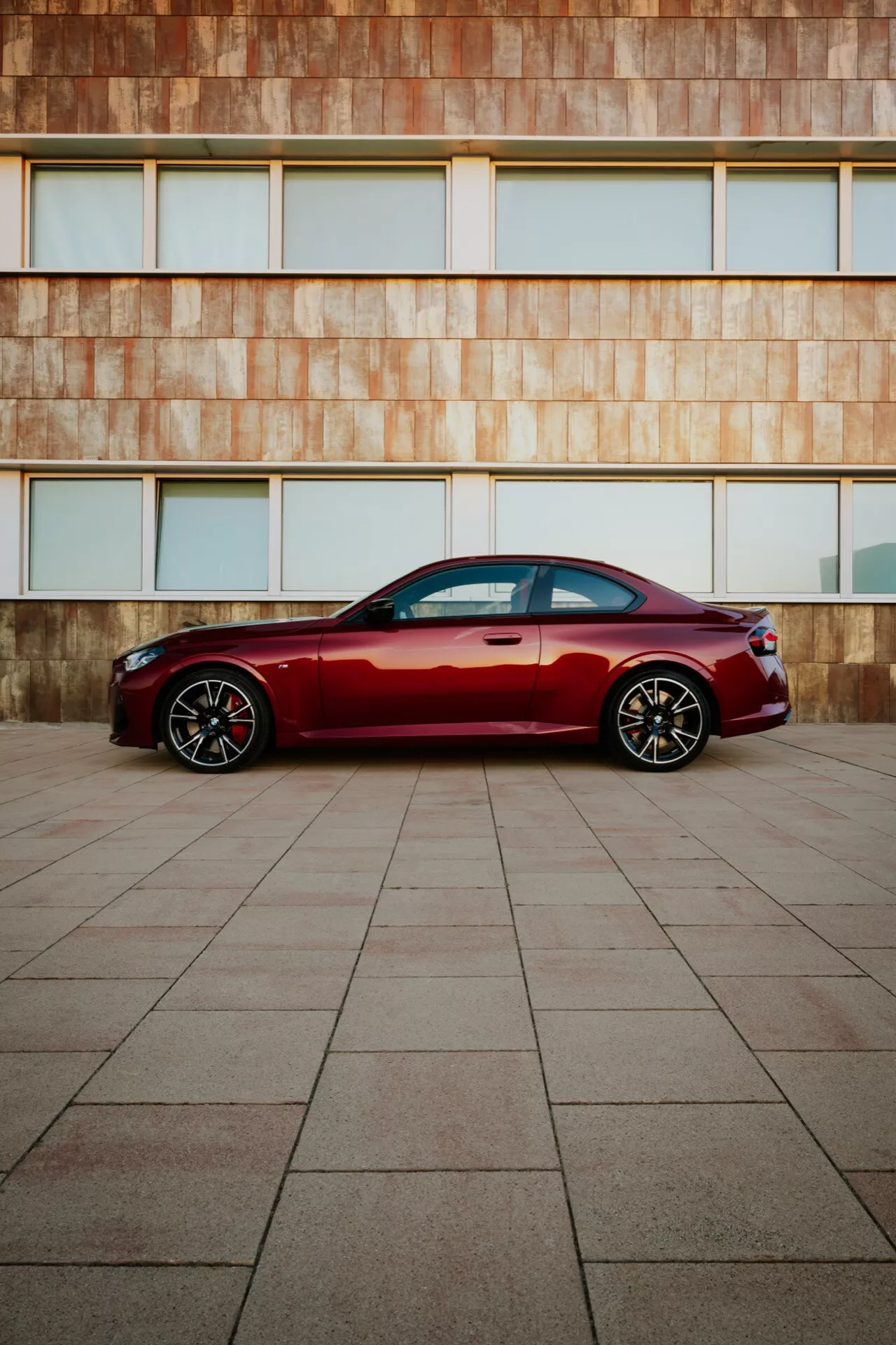 Modern lyxsedan djupt röd BMW 2er-Reihe 2024 (G42-G87 Facelift) parkerad på stor kaklad yta med rostfärgad arkitektonisk byggnadsfasad i bakgrunden, sidoprofilvy, elegant linjer, kromfönsterlister, sportiga aluminiumhjul, naturligt ljus.