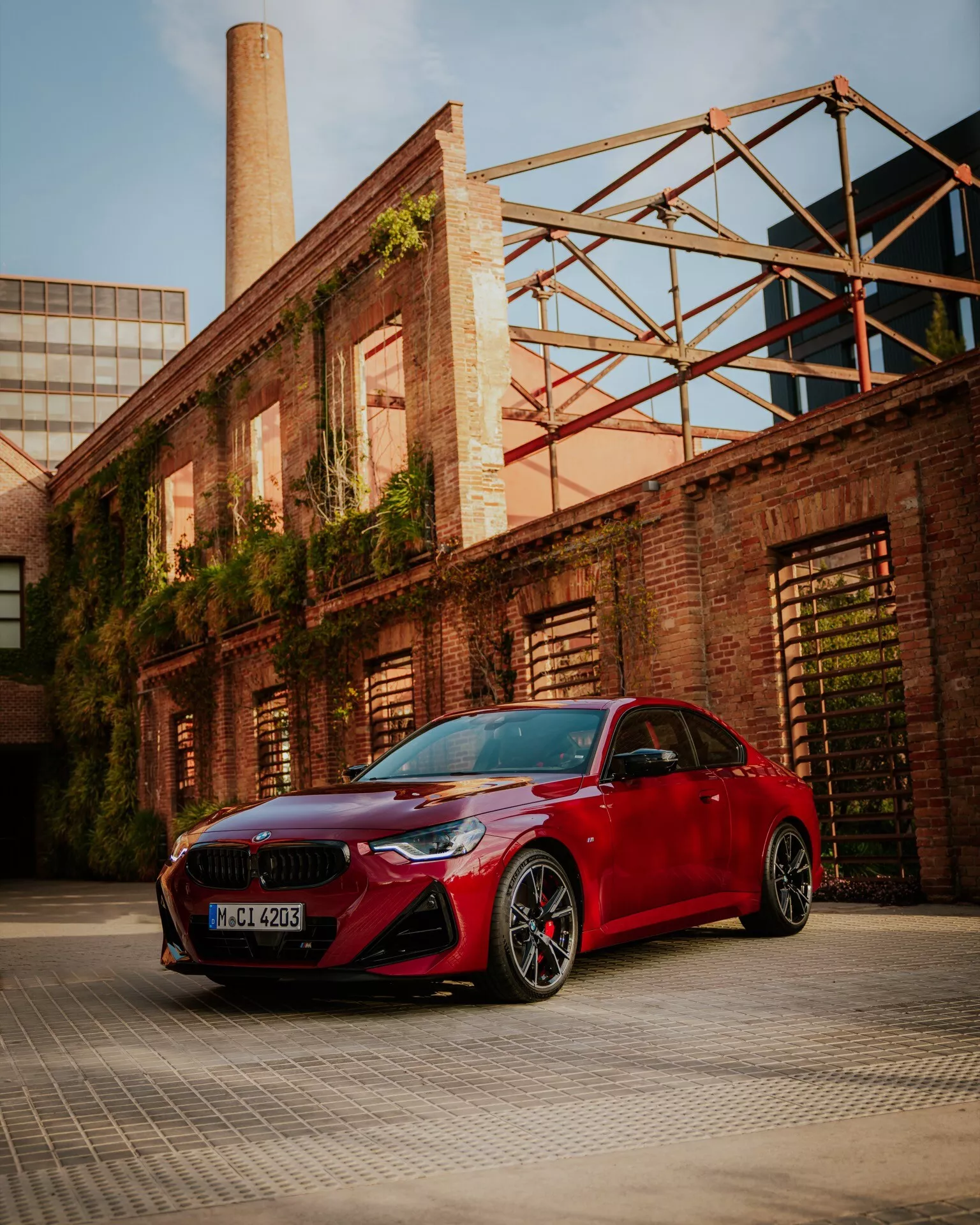 En låg vinkelbild av en röd BMW 2-serie 2024 (G42-G87-facelift) parkerad i ett industri- eller lagerdistrikt, som visar dess sportiga främre stötfångare med stora luftintag, smala eleganta strålkastare, njurformad grill och aerodynamiska sidospeglar. Bilden visar även flerekrade svarta lättmetallfälgar med lågprofildäck och synliga bromsok. De skulpterade linjerna på huven och den glänsande lacken framhävs av varmt omgivningsljus, vilket betonar bilens lyxiga och prestandainriktade designelement.
