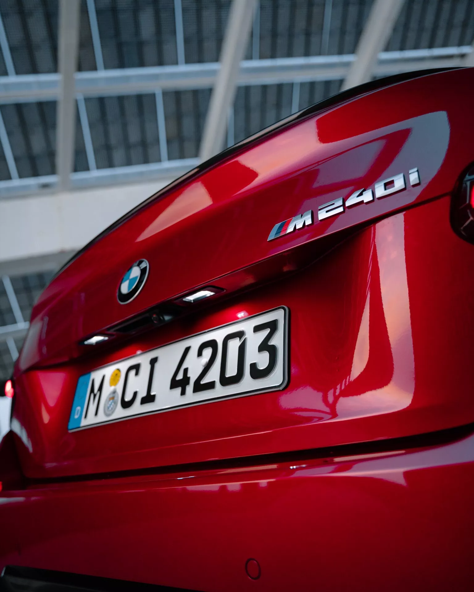 Närbild av bakpartiet på en röd BMW M240i, som visar BMW-logotyp, modellbeteckning, baklucka, L-formade baklyktor och integrerad bakspoiler, med en urban eller industriell bakgrund.