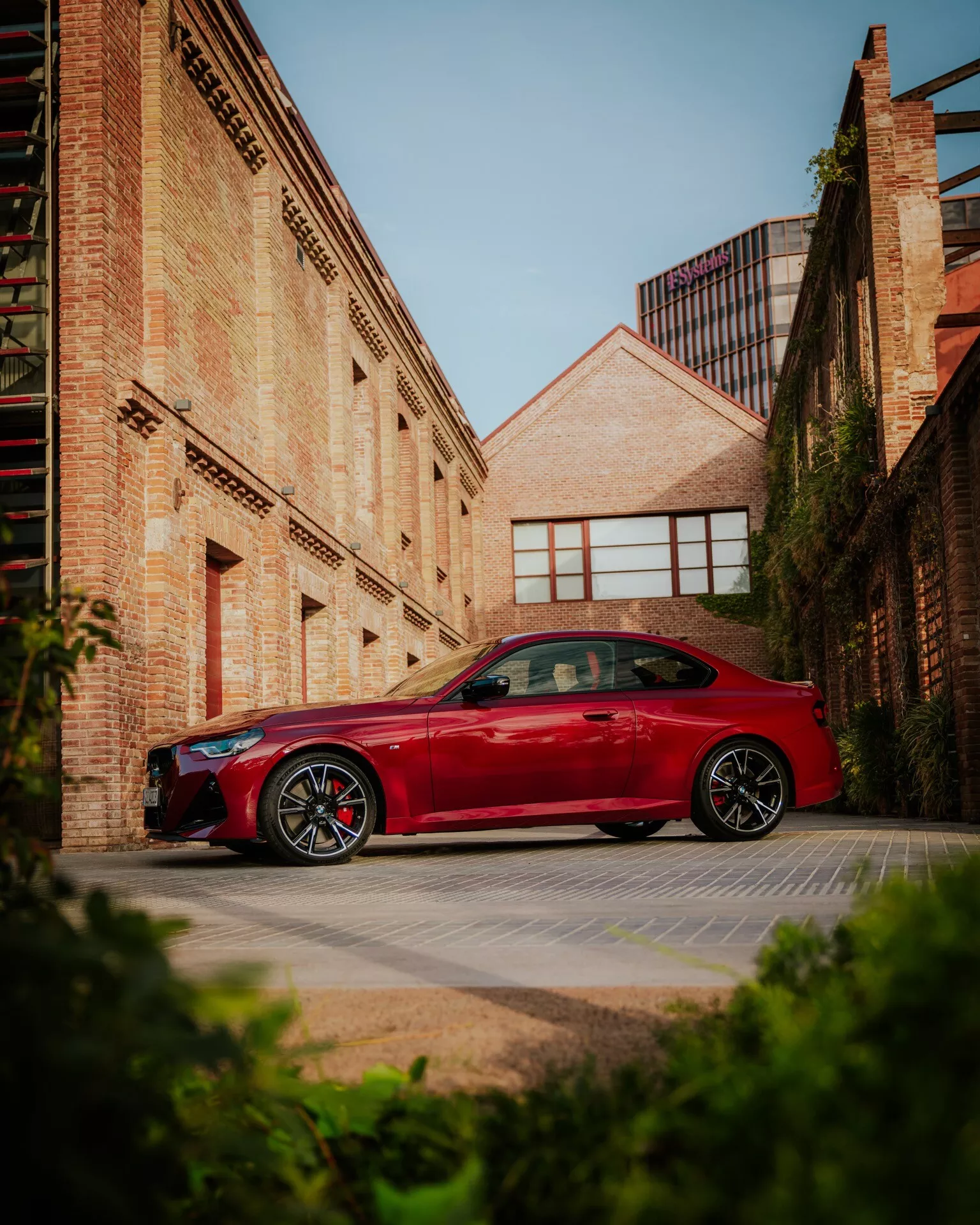 En röd BMW 2-serie 2024 (G42-G87 facelift) parkerad i en urban miljö, med tegelbyggnader i bakgrunden. Bilen visas i profilvy med designlinjer över dörrarna och skärmarna och tydligt synliga sportiga lättmetallfälgar. Bilden är tagen från en låg vinkel, vilket förstärker fordonets imponerande hållning mot morgon- eller eftermiddagssolen som kastar mjuka skuggor. Detta foto fångar teman om bil-design, sportiga detaljer och urban livsstil.
