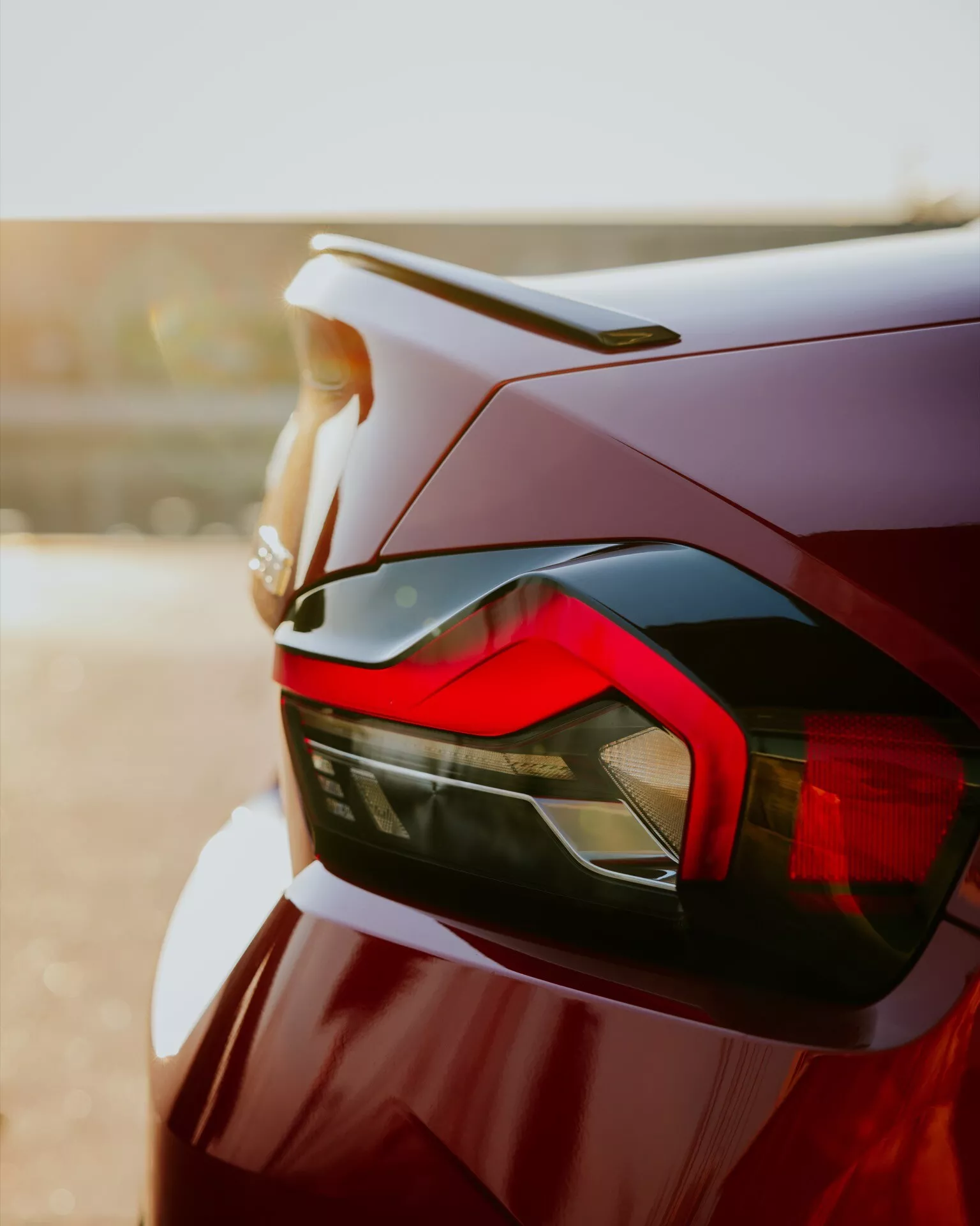 Närbild av bakdelen på en BMW 2-serie 2024 (G42-G87 facelift), som betonar fordonets designelement såsom den eleganta baklyktan med kantiga LED-signaturer, en subtil bakspoiler integrerad i bagageluckan och en högkvalitativ färgfinish i djupröd eller vinröd. Bilden, tagen från låg vinkel, framhäver bilens aerodynamik och moderna estetik, med en bokeh-bakgrund som skapar ett varmt sken.