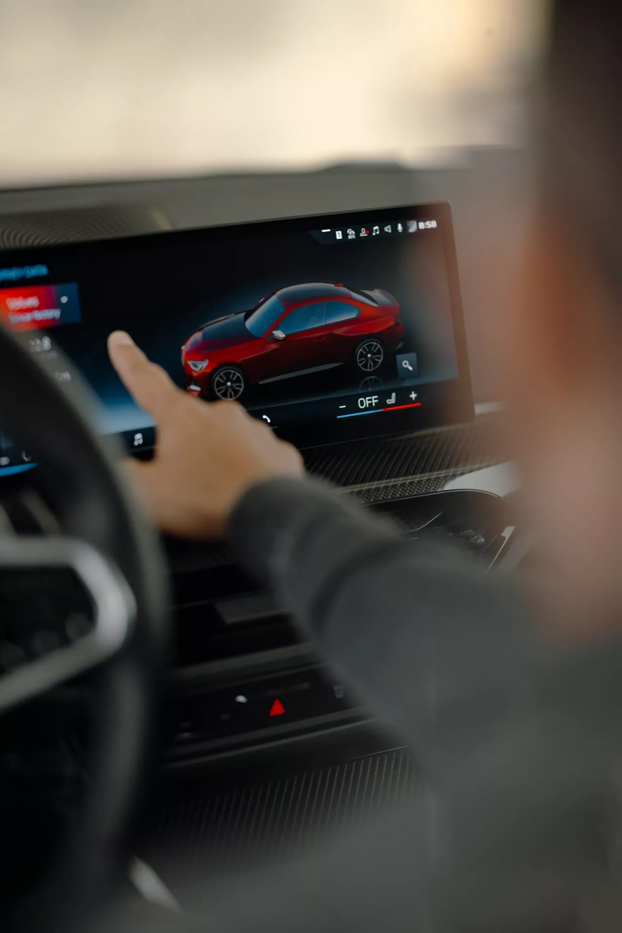 Närbild av ett modernt infotainmentsystem med pekskärm i bilen sett från förarsätet, med fokus på ett anpassningsgränssnitt som visar en röd sportbil. Ratten är något suddig vilket framhäver den avancerade tekniska uppsättningen av 2024 BMW 2-serien G42-G87 facelift med avancerade funktioner för navigation, inställningar, anslutning och multimedieunderhållning.