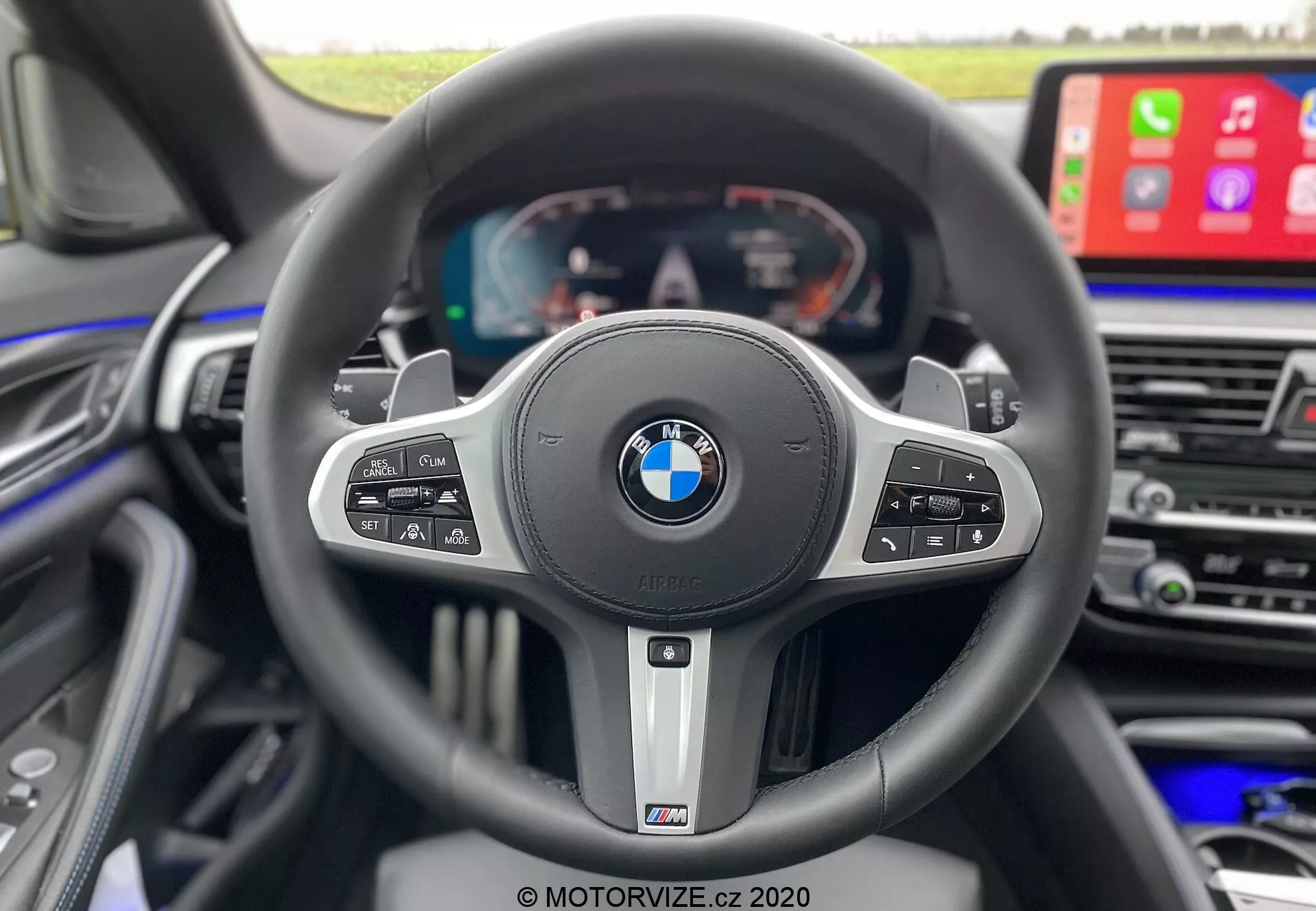 Interiörvy från förarens perspektiv inuti en BMW 520i Touring Steptronic från 2020, som framhäver en sportig treekrad ratt med BMW- och M-emblem. Ratten har flera kontrollknappar för farthållare, media, röstkommando och telefonfunktioner, samt växelpaddlar som indikerar automatväxellåda med manuella alternativ. Bakom ratten finns en helt digital display som visar hastighetsmätare, varvräknare och fordonsinformation. Till höger syns en del av mittkonsolen med en stor infotainmentskärm som visar Apple CarPlay, vilket betonar lyx, avancerad teknik och förarfokuserad design.
