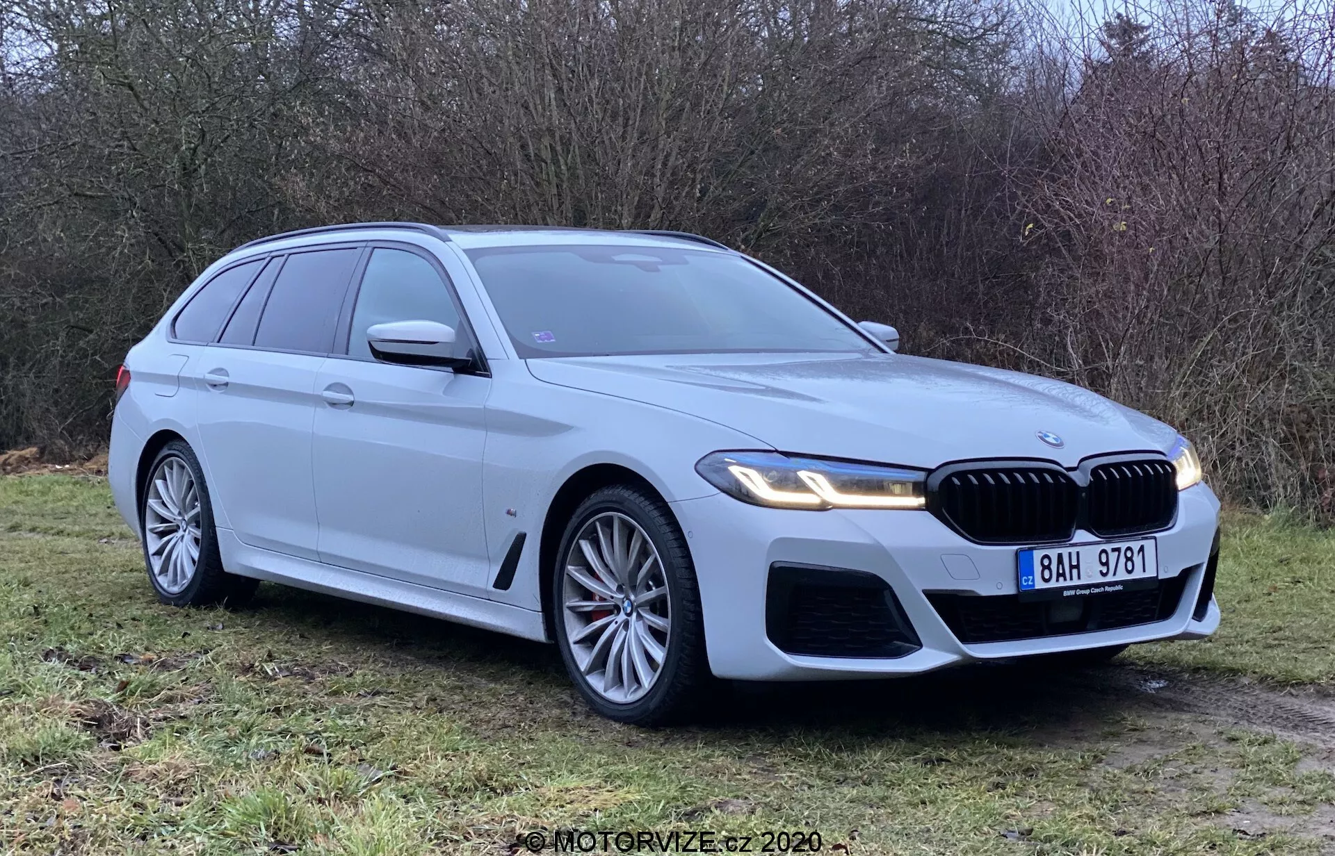 Silver BMW 5 Series Touring 2020 parkerad på gräs, sedd från ett trekvart frontvinkel som visar frontgrill, strålkastare och aerodynamisk design. Har LED-strålkastare, stora lättmetallfälgar, njurgrill och parkeringssensorer. Ren och ny, med betoning på BMW-märke, integrerade blinkers, takräcken och hajfensantenn.