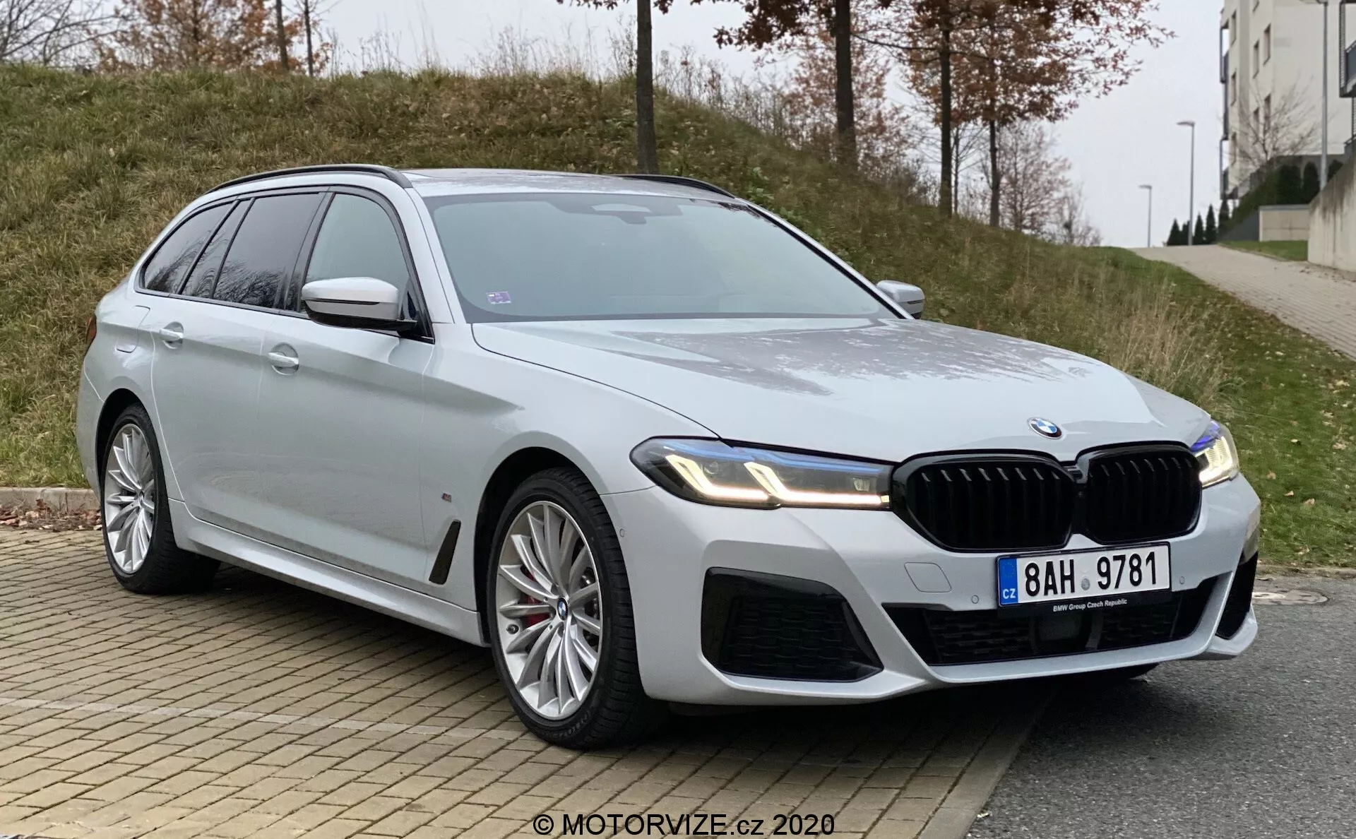 En modern och lyxig BMW 5-serie (BMW 520i Touring Steptronic) kombi fotograferad i en låg trekvarts vinkel framifrån. Bilden framhäver bilens främre grill, slanka vinklade strålkastare troligen utrustade med LED-teknologi samt de muskulösa designlinjerna på motorhuven och skärmarna. Synliga delar inkluderar frontgrill, stötfångare, strålkastare, förarsidans framdörr, framskärm, del av bakdörren, sidorutor, hjul och sidobackspegel på förarsidan. De stora lättmetallfälgarna antyder en sportigare variant av modellen och den silverfärgade lacken är reflekterande och ren. Bilen har en framträdande frontgrill med vertikala ribbor, ett märke på framskärmen som antyder en högre utrustningsnivå och sidospeglar med integrerade blinkers. Ett soltak är också synligt. Fordonet är parkerat vid en stadsväg med kantsten och trottoar i förgrunden samt ett gräsområde och en byggnad i bakgrunden, under molniga ljusförhållanden.