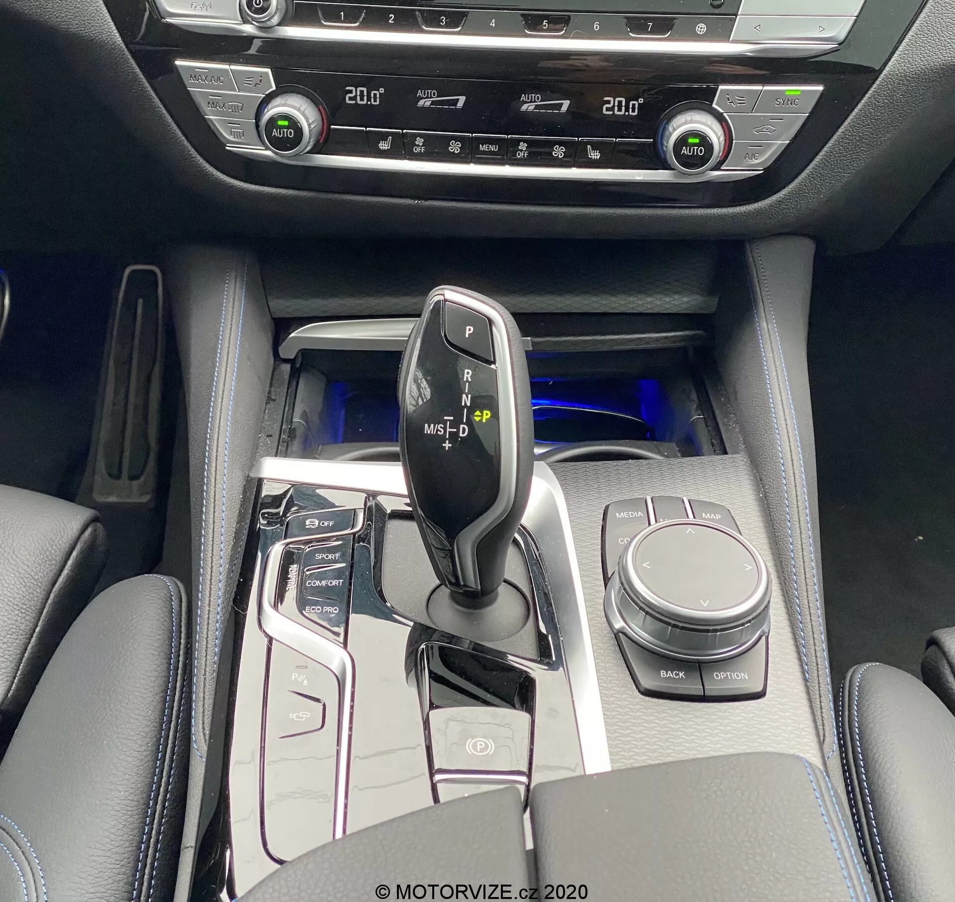 Fågelperspektiv av BMW 520i Touring Steptronic 2020 centralkonsol med en blank svart växelspak med metalliska accenter, omgiven av en högpolerad kontrollpanel med knappar för start/stopp-system, körlägen, parkeringssensorer och mer. Synliga är iDrive-kontrollvredet, mugghållare och vadderat armstöd med förvaring. Kallt blått ambient ljust förstärker känslan av lyx. Tvåzons klimatkontroll och sätesvärmare kontroller indikerar avancerade komfortfunktioner. Interiörmaterialen inkluderar läderliknande ytor med metalliska accenter och högpolerade paneler.