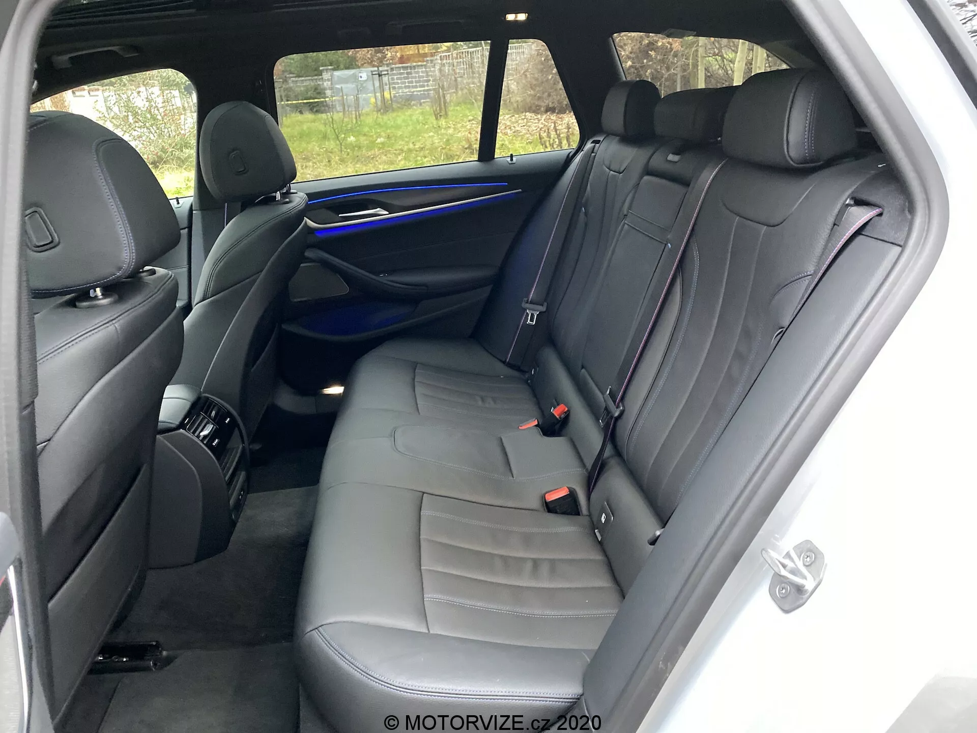 Interiören i en 2020 BMW 520i Touring Steptronic med fokus på baksätet från höger sida. Synvinkeln är nära den högra bakre passagerardörren och visar en bänk för tre med mörkt läderklädsel och accentstygn. Funktioner inkluderar tre huvudstöd, tre säkerhetsbälten, en dörrpanel med högtalargaller, handtag och kontrollfunktioner, en blå ambient belysningslist och ett förvaringsnät på baksidan av det främre passagerarsätet. Det välskötta interiören har inga synliga personliga föremål eller betydande slitage. Delvis utomhusvy genom bakrutan inkluderar ett staket.