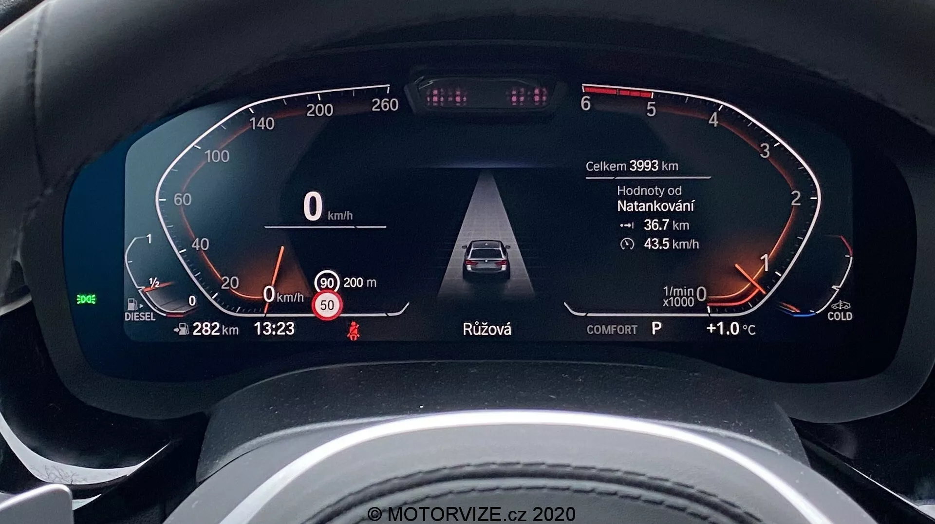 Närbild av den digitala instrumentpanelen i en BMW 5-serie 2020 (BMW 520i Touring Steptronic) tagen ur förarens perspektiv. Den digitala instrumentklustret visar nyckelinformation som fordonsfart (0 km/h), en bränslemätare för diesel, varningar för öppen dörr och säkerhetsbälte, avancerade förarassistanssystem som adaptiv farthållare, varvräknare i viloläge med en kall motorindikator och ytterligare data inklusive körsträcka (39933 km), utomhustemperatur (+1,0°C), tid (13:23), växelval (P för parkering) och körläge (komfort). Designen är elegant och modern, vilket indikerar ett exklusivt fordon.