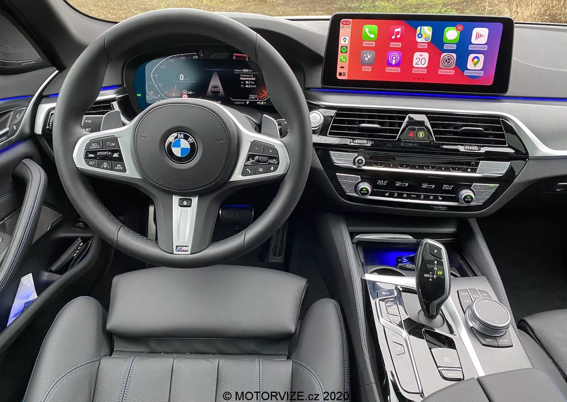 Interiörvy från förarsätet i en BMW 520i Touring Steptronic 2020, med en multifunktionell ratt i läder med BMW-logotyp och kontrollknappar för farthållare, ljud och telefoni. Den digitala instrumentpanelen visar hastighetsmätare, varvräknare och en navigationskarta. Mittkonsolen rymmer ett stort pekskärms infotainmentsystem som visar mobilappar som Apple CarPlay. Nedanför finns luftventiler, klimatkontrollpanel, ljudkontroller, en elegant designad växelspak och funktionsknappar. Interiören visar sittplatser med lädersöm, ambient belysning och högkvalitativa material med metalliska accenter.