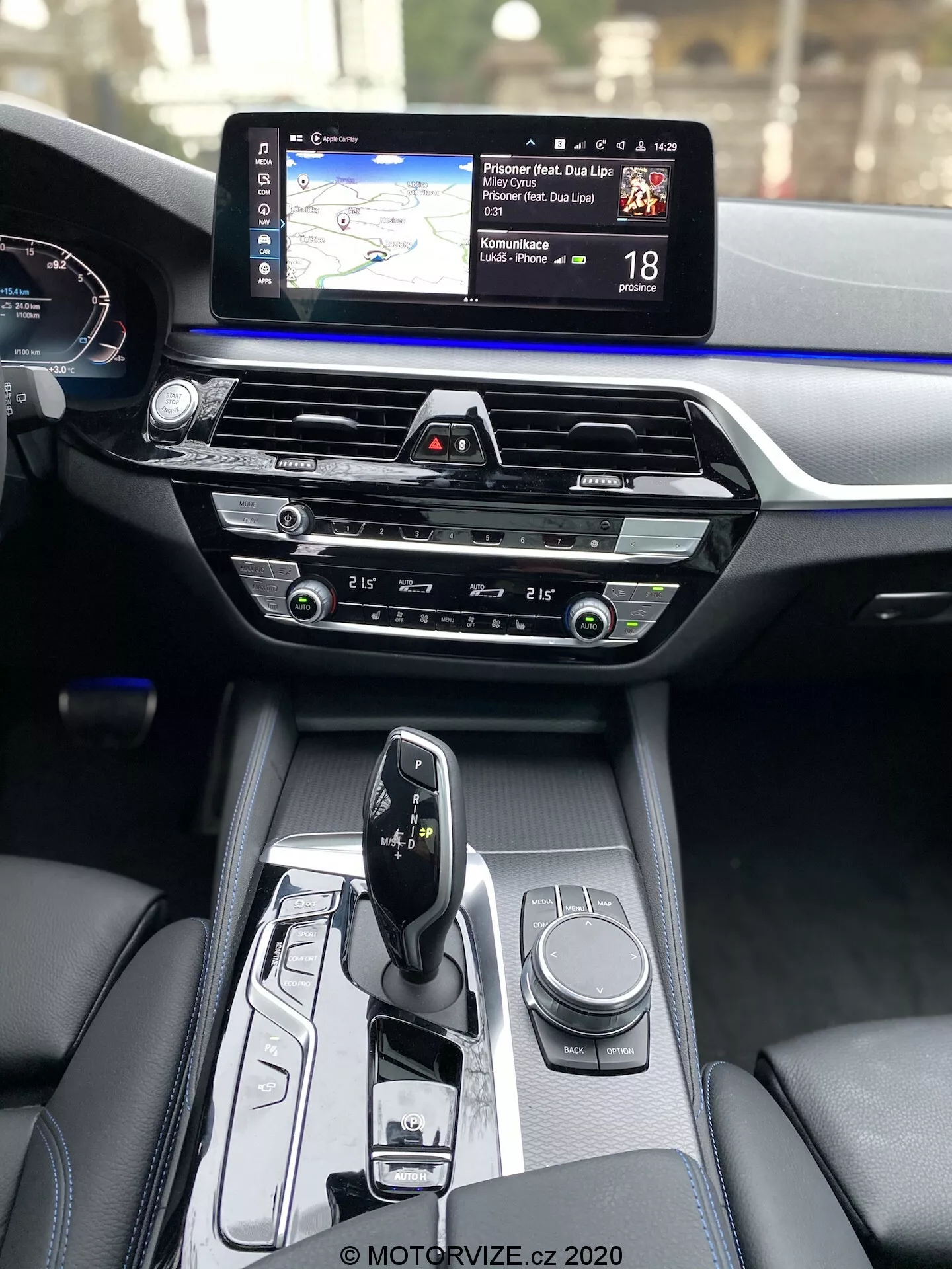 Interiörvy av BMW 5-serie 2020 (BMW 520i Touring Steptronic), som visar mittkonsolen och instrumentbrädan från främre passagerarens perspektiv. Mittkonsolen har en elegant automatisk växelväljare i 'park' läge, med olika kontroller inklusive en roterande ratt för infotainmentsystemet och knappar för körlägen eller fordonsinställningar. Mitten av instrumentbrädan har klimatkontroller med intuitiva rattar och knappar, ovanför dessa finns en CD-spelare. Överst på instrumentbrädan finns en bred infotainment pekskärm som visar navigationskarta och hastighetsbegränsningsinformation. Delvis synligt är det digitala instrumentklustret som visar hastighetsmätaren. Interiörmaterialen framhäver premiumfinish, med kontrast mellan mörkt läder och en blå accentlist över hela instrumentbrädans bredd.