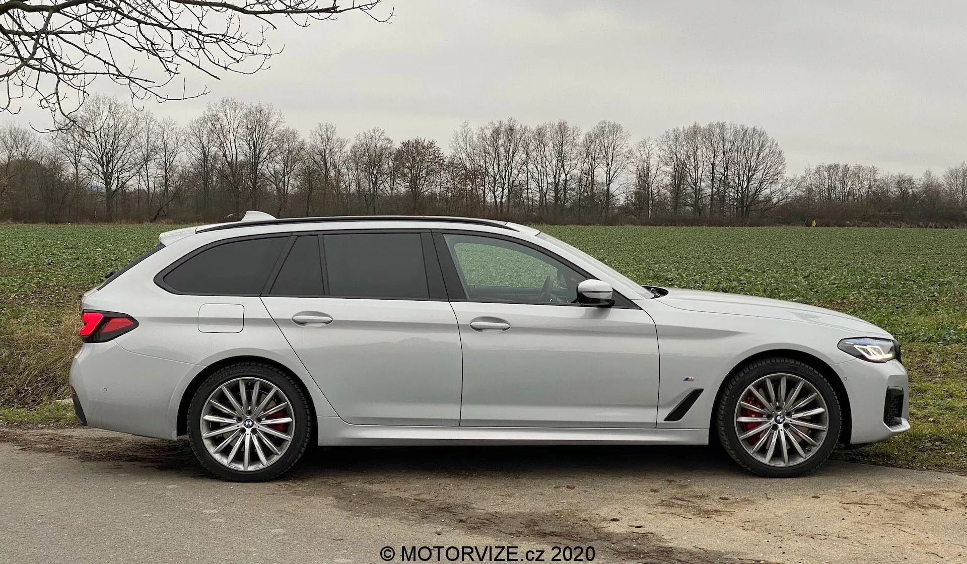 Sidprofil av en modern BMW 520i Touring Steptronic kombi tagen i dagsljus, som visar eleganta designlinjer, fem dörrar inklusive bakluckan, en lång taklinje med en takspoiler, stora mångekrade lättmetallfälgar med synliga bromsok, karossfärgade dörrhandtag, integrerade indikatorer i sidospeglarna, lätt tonade rutor och en matt eller icke-metallisk lackfinish. Bilen är placerad på en grus- eller jordkant bredvid en asfalterad väg i en lantlig miljö, med gräs och träd i bakgrunden, under gynnsamma väderförhållanden.