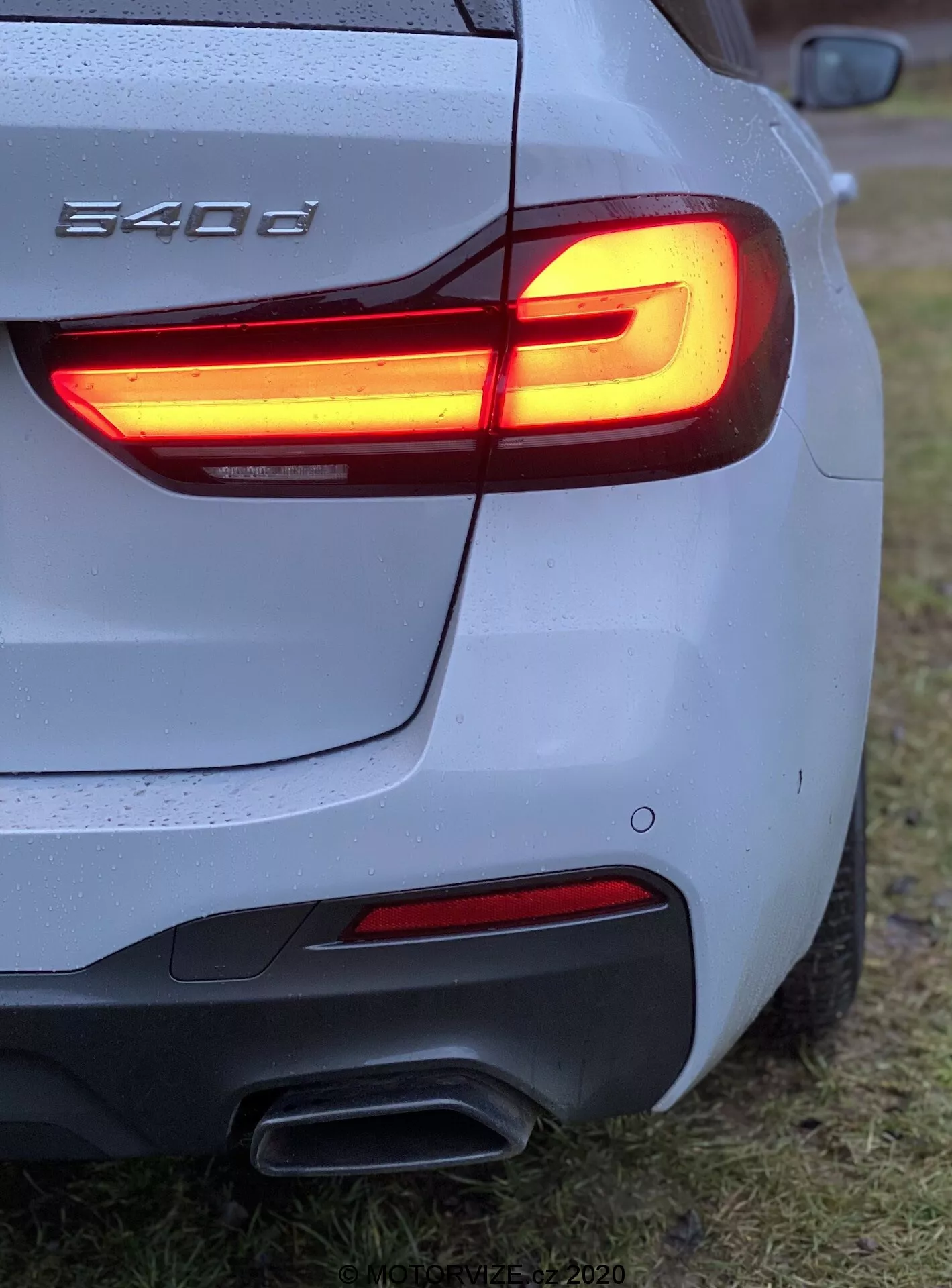 Närbild av bakre trebarnsvinkel av en BMW 540i, som visar L-formad LED-baklykta, dubbla avgasrör, silver/grå kaross med vattendroppar, skulpterad bakre stötfångare, delvis synlig BMW-märke, parkerad på gräsbevuxet område.