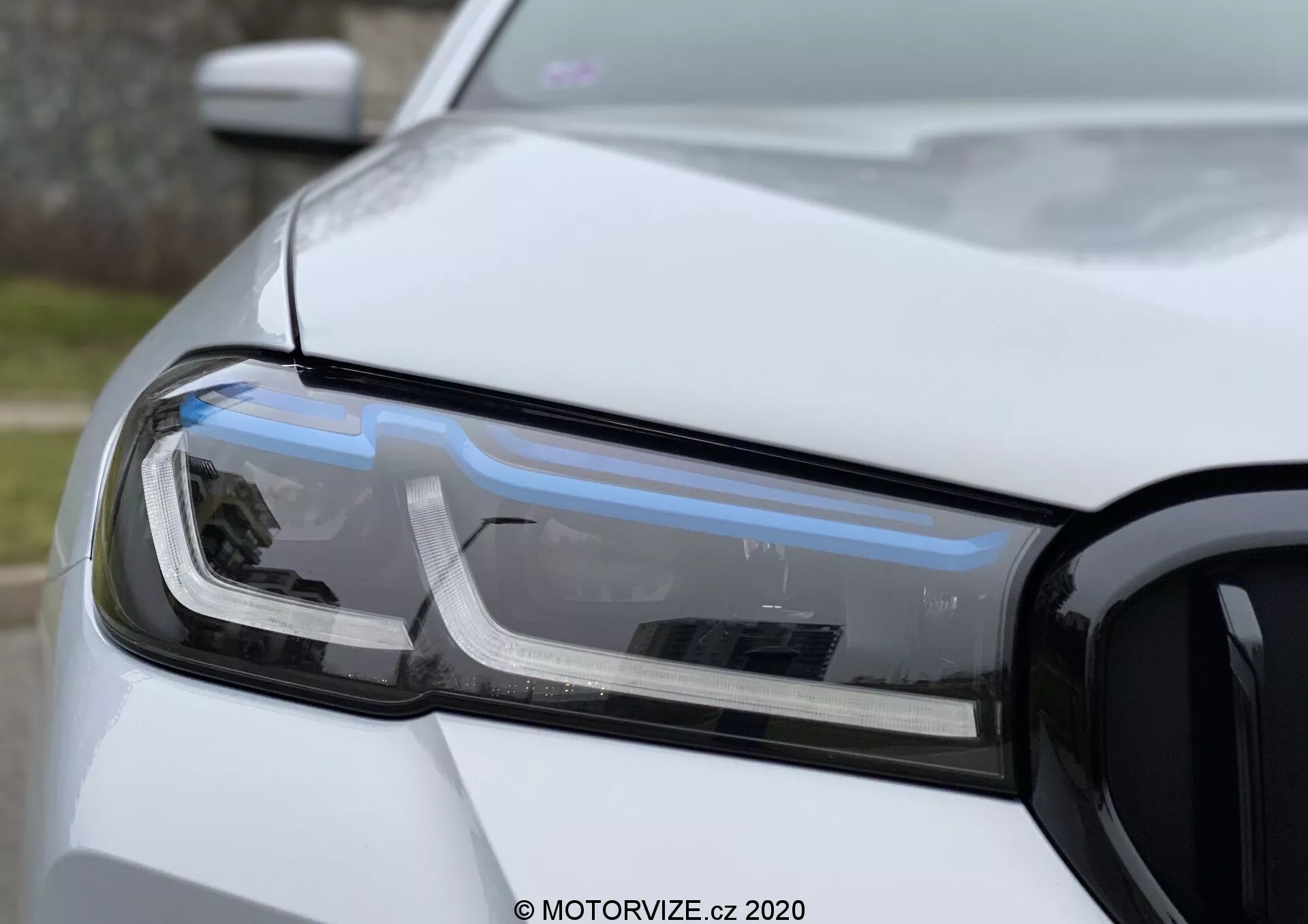 Närbild på förarsidans strålkastarenhet på en BMW 520i Touring Steptronic, med fokus på LED-dagljus, projektorlampa och blinkers. Fotot visar bilens vänstra främre sektion, inklusive delar av huven, främre stötfångaren och skärmen, med grillen synlig till höger. Bilden har ett grunt skärpedjup som framhäver strålkastarens teknik och design i naturligt, diffust ljus med en solid, icke-metallisk vit lack.