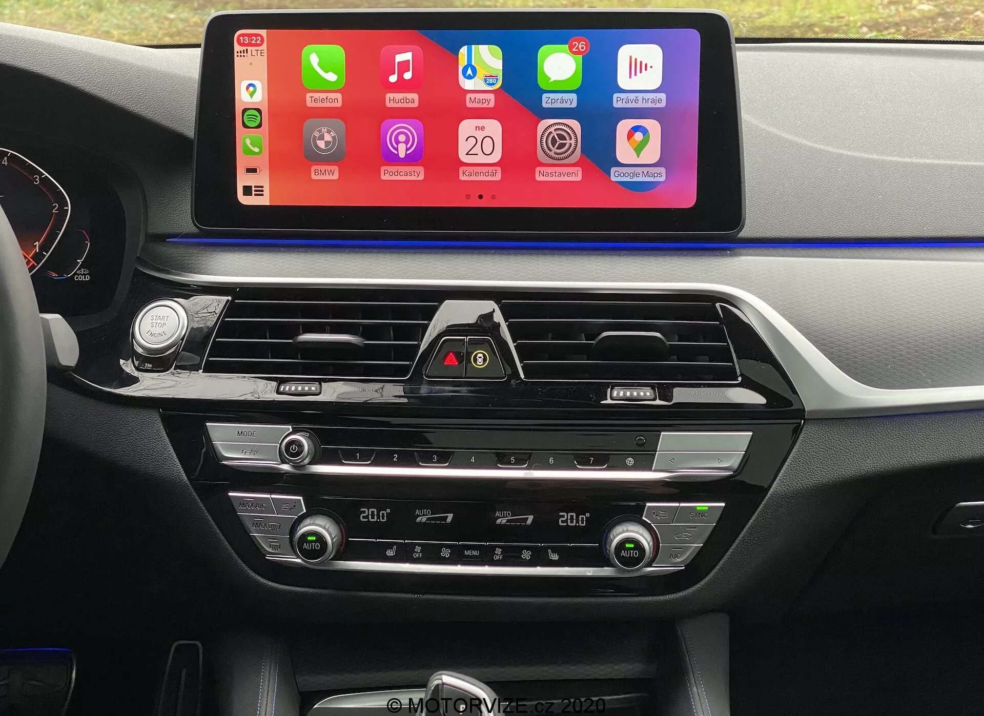 Interiorsbild av en BMW 5er-Reihe 2020 (BMW 520i Touring Steptronic) från förarsätets perspektiv, med fokus på den centrala instrumentbrädan. Det mest framträdande inslaget är en stor, horisontellt orienterad infotainmentskärm med färgglatt gränssnitt som stöder smartphone-integration som Apple CarPlay och Android Auto. Under skärmen finns luftventiler med kromdetaljer och en varningsljusknapp. Längre ner finns tvåzons klimatreglage med digitala displayer för temperaturinställningar samt knappar och rattar för justering av lufttemperatur, fläkthastighet och luftdistribution. Instrumentbrädan och mittkonsolen har mjuk plast och metallföremål, vilket ökar den estetiska attraktionskraften.