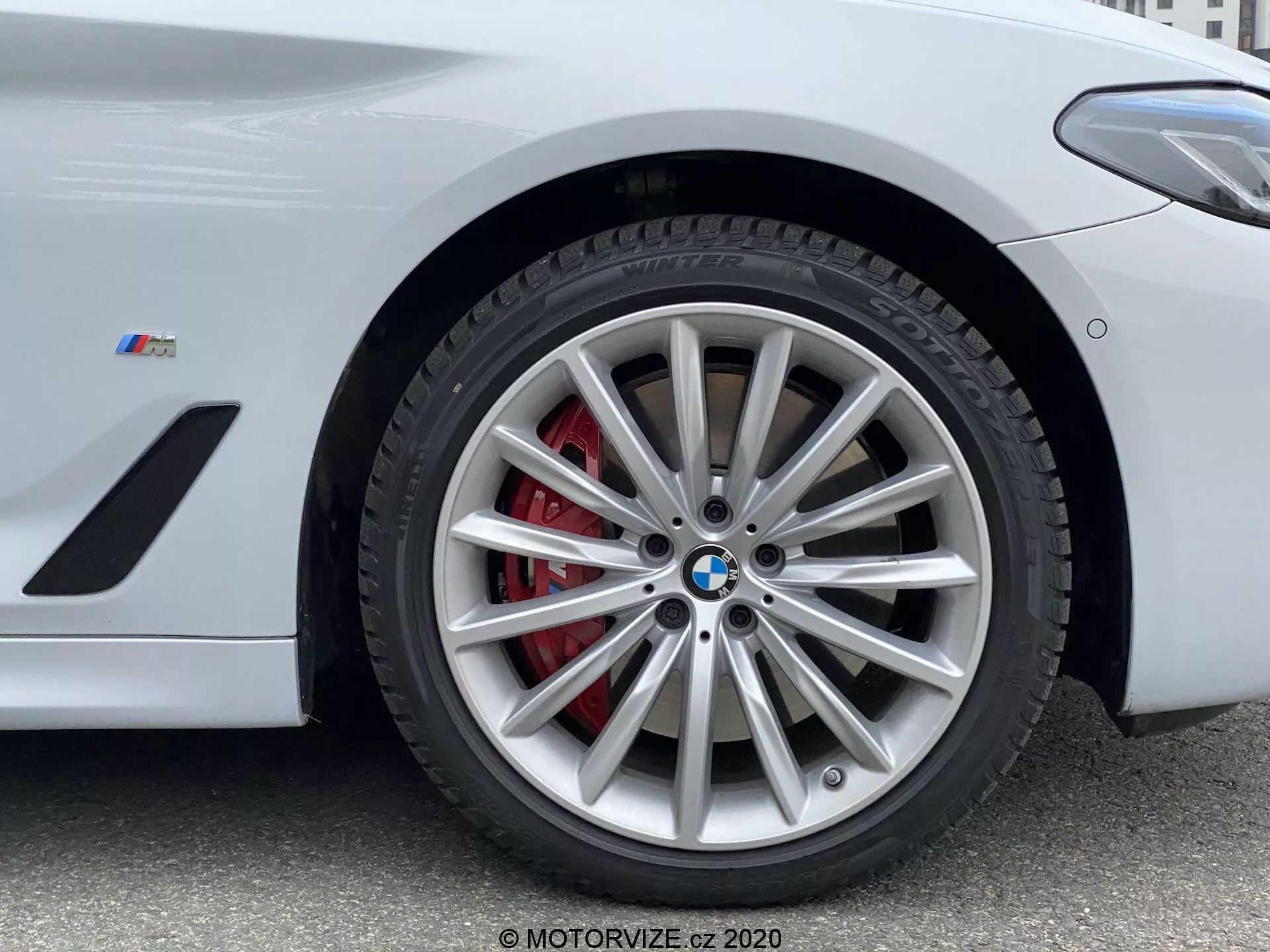 Närbild av framhjulssektionen på en BMW från 2020 års 5-serie, specifikt modellen 520i Touring Steptronic. Fotot taget från låg vinkel framhäver fälgens intrikata flerspakiga design med silverfinish, kompletterad med ett rött bromsok och en ventilerad rotor. BMW-emblemet och 'M'-märket på skärmen antyder att detta är ett prestandaorienterat fordon från BMW:s M-serie. Bilden fångar nyckeldetaljer som är lämpliga för att visa högpresterande bromssystem, lyxig fordonsdesign och däckteknik.