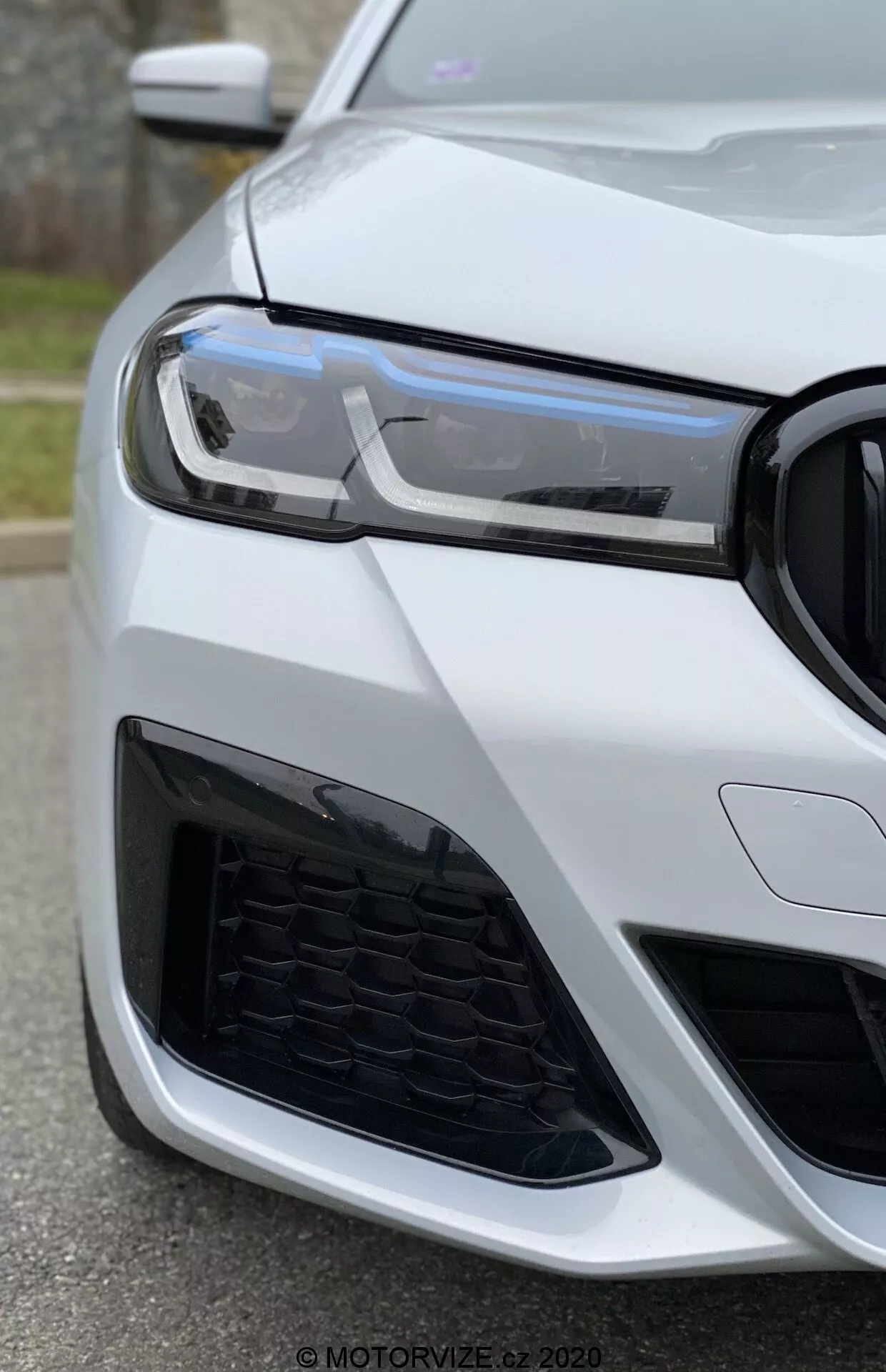 Närbild på fronten av en BMW 520i Touring Steptronic (2020) taget från en låg vinkel med fokus på strålkastaren, grillen och stötfångaren. Moderna LED-dagljus, honungskakemönster i grillen och den aggressiva luftintagsdesignen antyder en sportig och avancerad bil. Bilen är målad i glansigt vitt som framhäver dess lyxiga och välskötta utseende.