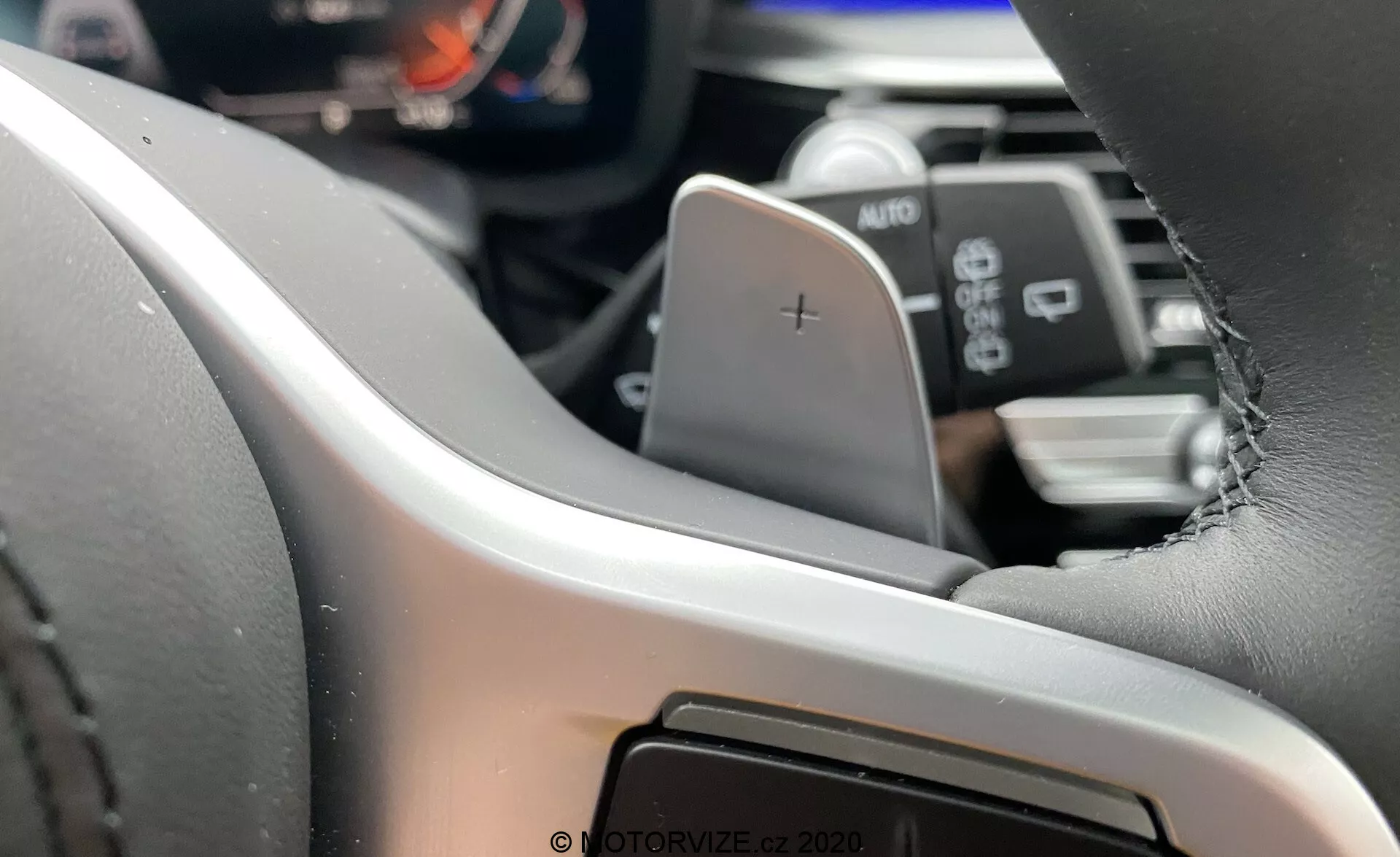 Interiörvy av BMW 5-serien 2020 (BMW 520i Touring Steptronic) med fokus på vänster sida av ratten från förarens perspektiv⁠—modern ratt klädd i läder med synliga sömmar, utrustad med en uppväxlingspaddel markerad med '+'. Den digitala instrumentbrädan eller instrumentpanelen är delvis synlig, vilket belyser avancerade informationssystem för föraren. Under paddeln finns en spak märkt AUTO, vilket troligtvis indikerar strålkastare eller vindrutetorkarkontroller. I den suddiga bakgrunden syns en antydan om infotainmentsystemet eller centralkonsolen, vilket antyder en pekskärmsgränssnitt.