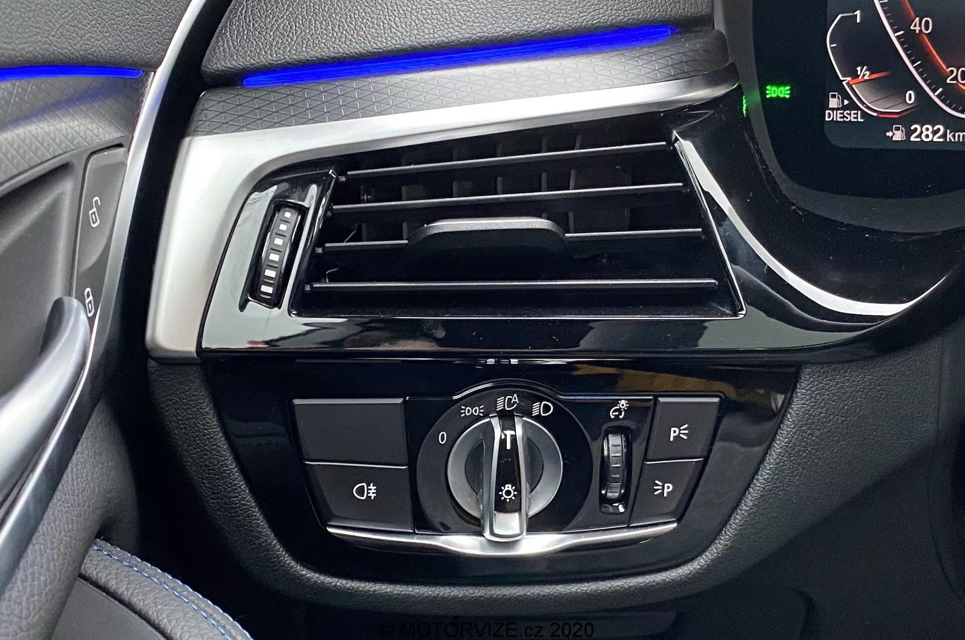 Närbild av interiören i BMW 5-serien 2020, med fokus på förarsidans instrumentpanel med en blå ambient stripe ovanför den horisontella ventilen. De centrala kontrollerna inkluderar en roterande ratt för automatiska och strålkastarinställningar, knappar förmodligen för dimljus och strålkastarnivåering, ytterligare förarassistansknappar och en digital hastighetsmätare som visar 0 km/h med bilen i parkeringsläge och 282 km på mätaren. En 'DIESEL'-indikator är också synlig.