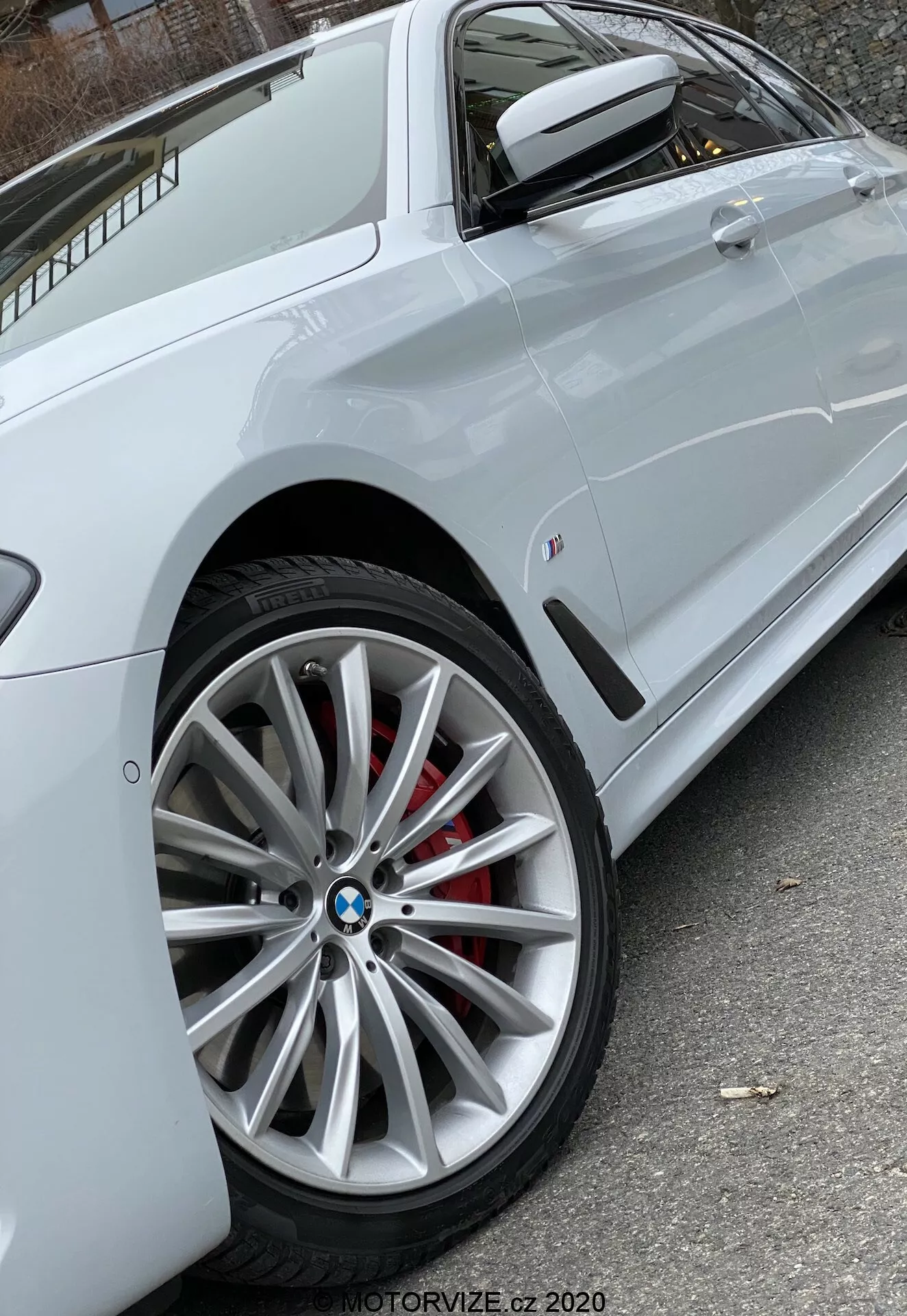 Närbild från sidan på en vit BMW 520i Touring Steptronic (2020), fokuserad på den moderna flerspåriga legerade framhjulet med synlig BMW-logotyp och röd bromsok, delvis visande sidoprofil med skarp karaktärslinje genom dörrhandtagen, färgmatchad sidospegel med inbyggd blinkers och en liten badge på den främre sidopanelen. Det naturliga ljuset säkerställer tydlig synlighet av fordonets egenskaper. Foto av MOTORVIZE.cz 2020.