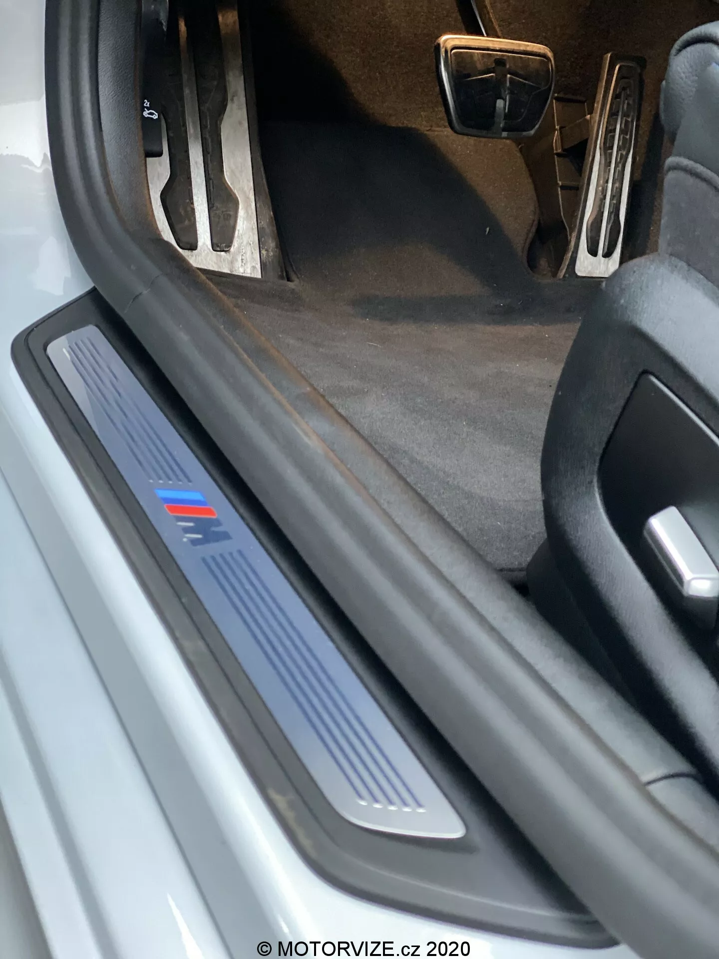 Närbild av förarsidans tröskel till en BMW 520i Touring Steptronic 2020. Bilden visar ett lågvinkelperspektiv och framhäver tröskelplattan i borstad metall med en blå rand, som kan indikera en specifik utrustningsnivå eller sportutgåva. Den öppna dörren är inte synlig, men den skyddande och dekorativa tröskelplattan är tydlig, vilket antyder möjlig LED-belysning. Den mörka interiörmattan och den delvis synliga metallfinishen på pedalerna indikerar ett möjligt sportigt interiörpaket. Synligt skräp och slitage på tröskelplattan antyder att bilen är begagnad.