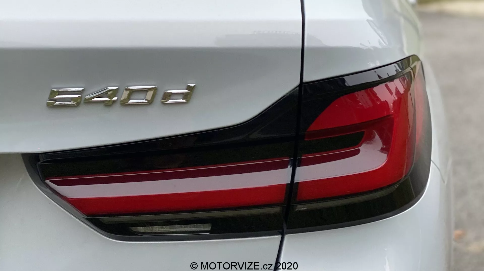 Närbild av den bakre delen av en BMW 520i Touring Steptronic, med fokus på vänster baklykta och en del av bagageluckan. Vinkeln är något sned från bakre vänstra hörnet och framhäver den moderna LED-baklyktan med sin skarpa, kantiga design och framträdande röda sektion. Bagageluckan visar modellbeteckningen '840d.' Karossen är glansig, troligen vit eller ljus silver, vilket framhäver lyx och precisionsingenjörskonst.