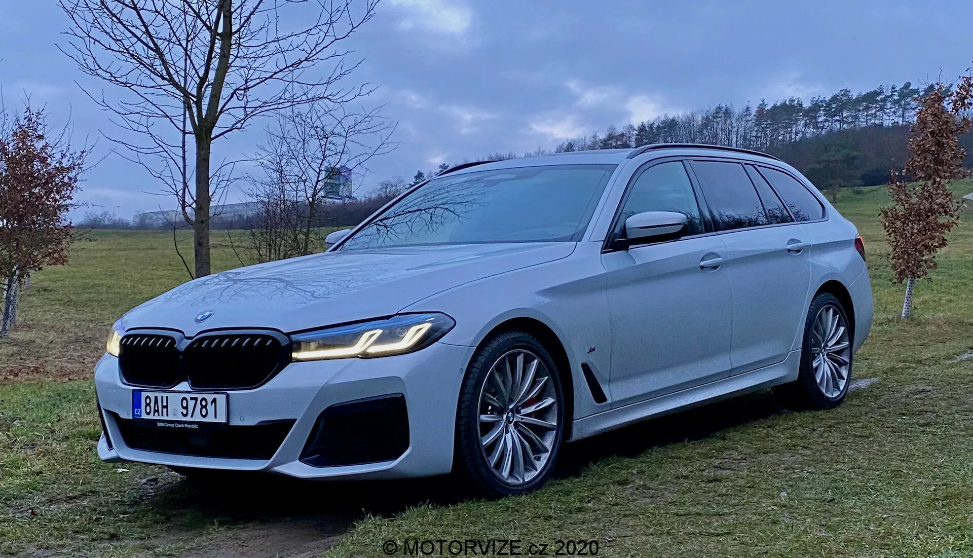 Bild av en 2020 BMW 5-serie Touring (BMW 520i Touring Steptronic) lyxkombi i ljus silver eller grå färg, som visar dess aggressiva och sportiga front med stora luftintag, framträdande grill och eleganta LED-strålkastare. Fordonet har stora lättmetallfälgar med flerradie-design och noterbara bromsok, en lång taklinje med takspoiler, kromade fönsterlister och stark skuldralinje. Fotograferad från en medelhög perspektiv i en gräsbevuxen, halvlandlig miljö med diffust ljus.
