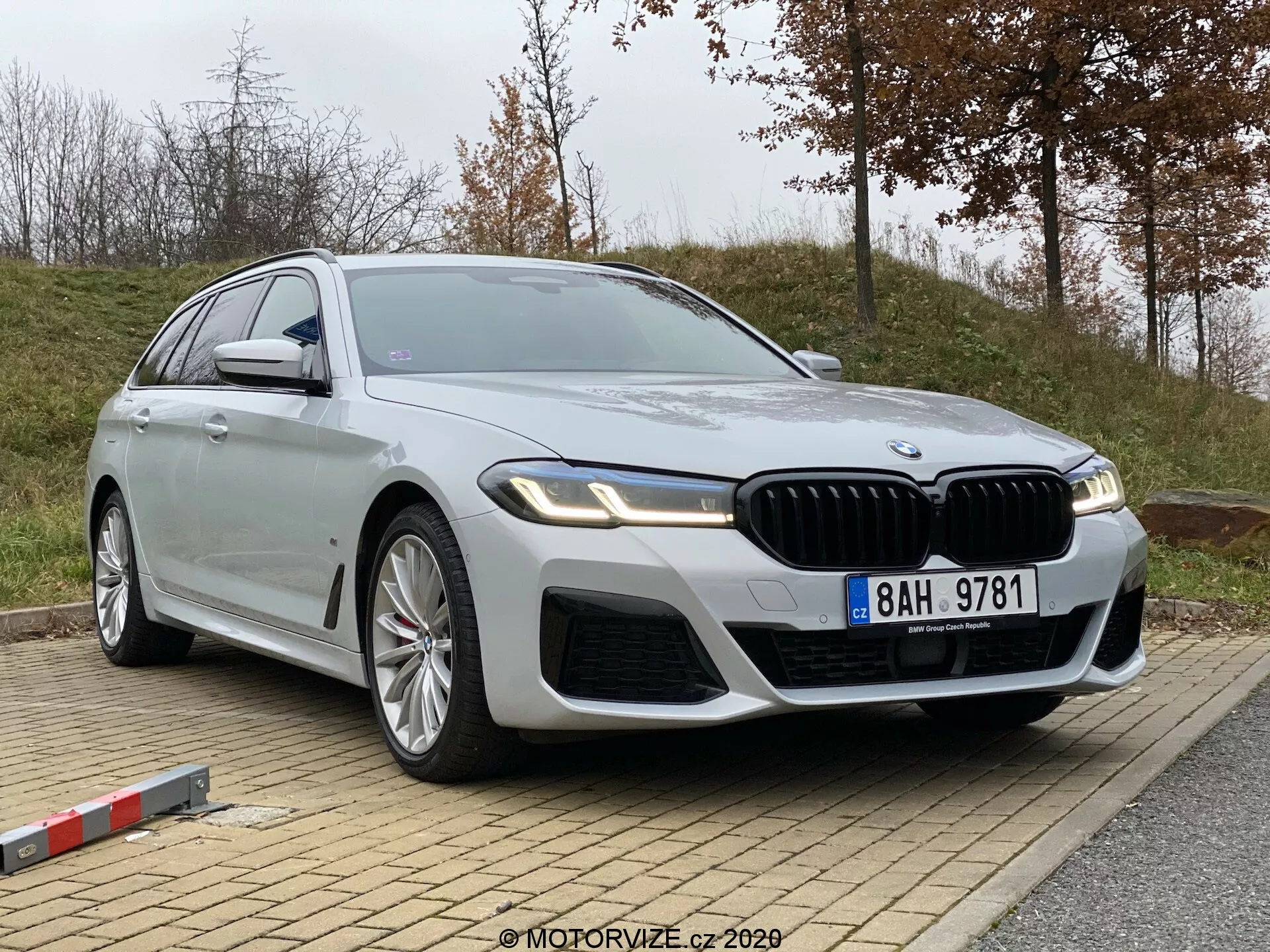 Bilden visar BMW 5-serien 2020 (520i Touring Steptronic), en modern lyxsportbil, parkerad på en lätt sluttning. Fotot är taget från en låg och något framifrån avvinklad vinkel, vilket lyfter fram fordonets främre kvartsdel och profil. Noterbara detaljer inkluderar ett framträdande njurformade galler, intrikata luftintag på den främre stötfångaren, eleganta, vinklade LED-strålkastare, stora flerstrålade lättmetallfälgar, en lång svepande karosssiluett med metallic silvrig finish, kromdetaljer, integrerade blinkers i sidospeglarna och potentiellt avancerad teknik som autonoma körassistenter och parkeringsassistans. En badge är synlig på den främre kvartspanelen, vilket indikerar en specifik modellvariant.