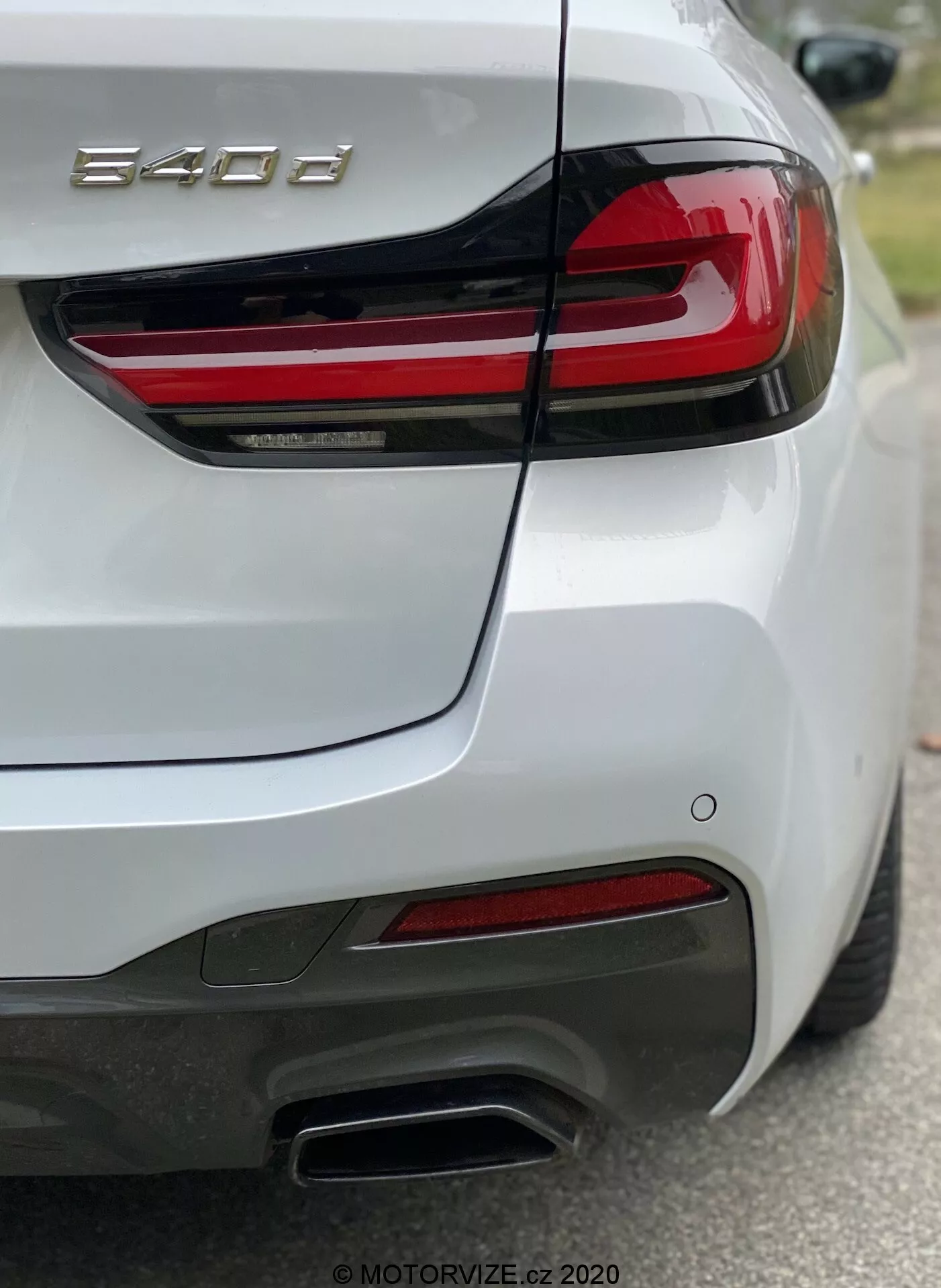 Bakdelen av en BMW 8-serie (840i) personbil, fotograferad från bakre högra kvartalssynvinkel, framhäver det högra bakljuset med horisontella röda LED-remsor, formad bagagelucka med integrerad spoiler, bakre stötfångare med sensorer, dubbla metalliska avgasrör på högersidan, röda reflexer och vit blank lackering. Fotografiet fokuserar på '840i' -märket, visar detaljerade design- och tekniska funktioner som belysning och förarassistanssystem.
