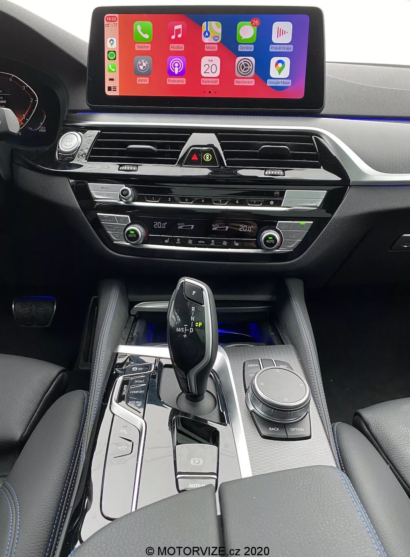 Interiör bild av en 2020 BMW 520i Touring med Steptronic transmission, fotograferad från förarens perspektiv och lätt förhöjd för att ge en tydligare vy av mittkonsolen, växelspaken och en del av instrumentbrädan. Bilden visar en stor infotainmentskärm som visar en anslutningsinterface för smartphones, troligen Apple CarPlay, monterad högt på instrumentbrädan för enkel synlighet. Nedanför skärmen finns moderna, horisontella luftventiler med kromdetaljer, följt av taktila klimatanläggningsknappar och rattar. Mittkonsolen innehåller också kontroller för ljudsystemet, ytterligare klimatanläggningskontroller och eventuellt en CD-spelarslot. Den ergonomiska och minimalistiska växelspaken, troligen elektronisk, är omgiven av svart högblank trim och metalliska accenter, med knappar för körlägesval och en navigationsvred. Interiören innehåller också armstöd klädda i läder med synliga sömmar, vilket ger en lyxig känsla. Mjuk vit belysning markerar knappar runt växelspaken, vilket indikerar en automatisk växellåda.