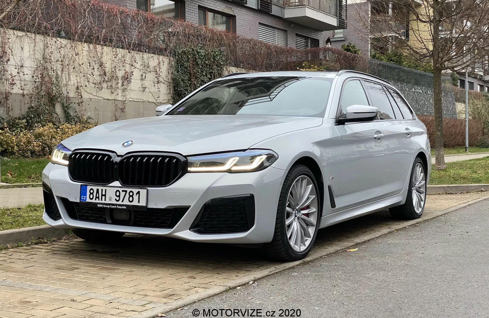Lyxig BMW 520i Touring parkerad på en stadsgata, sedd från en låg frontal trekvartsvinkel som betonar bilens framsida och förarsida. Bilen har en framträdande njurgrill med kromdetaljer, eleganta LED-strålkastare, sportig främre stötfångare och definierade veck på motorhuven. Mångekrade lättmetallfälgar, sidobackspeglar med blinkers och kamera, krominramning av fönstren och ett soltak bidrar till ett lyxigt utseende. Placerad på höger sida med gräs, buskar och bostadshus i bakgrunden, vilket antyder avancerad teknik och högkvalitativ design.