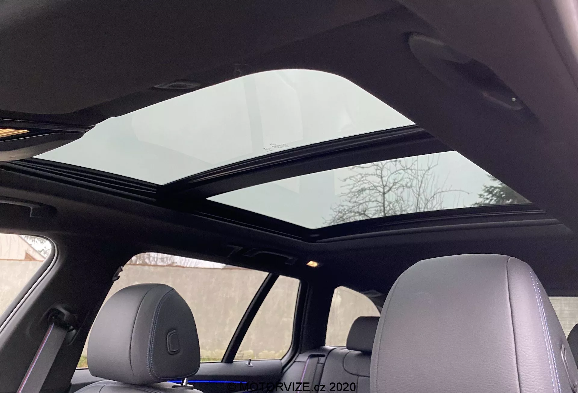 Inrikesvy av en BMW 5-serie 2020 520i Touring med Steptronic-transmission som visar ett panoramasoltak som sträcker sig över fram- och baksätet från perspektivet av en passagerare i baksätet som tittar uppåt. Panoramasoltakets främre panel är något lutad öppen, vilket förbättrar ventilationen. Taktäckningen är ljusfärgad och kompletterar bilens sammanhängande inredningsdesign, med nackstöd som matchar interiörmaterialet och framhäver avancerad komfort och moderna bekvämligheter.