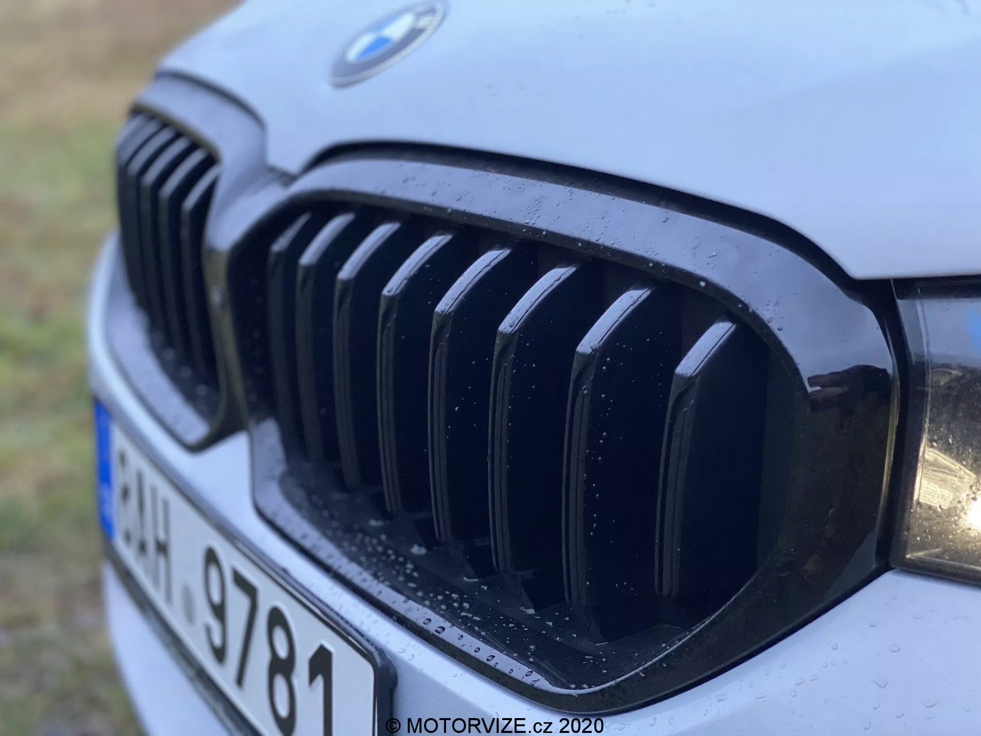 Närbild av frontgrillen på en BMW 520i Touring Steptronic från modellår 2020, som visar den karakteristiska njurgrillen och BMW-emblemet med vertikala svarta slattar, delvis strålkastare och vattendroppar som antyder nyligen regn eller tvätt, tagen från en låg framsida-vänster vinkel.