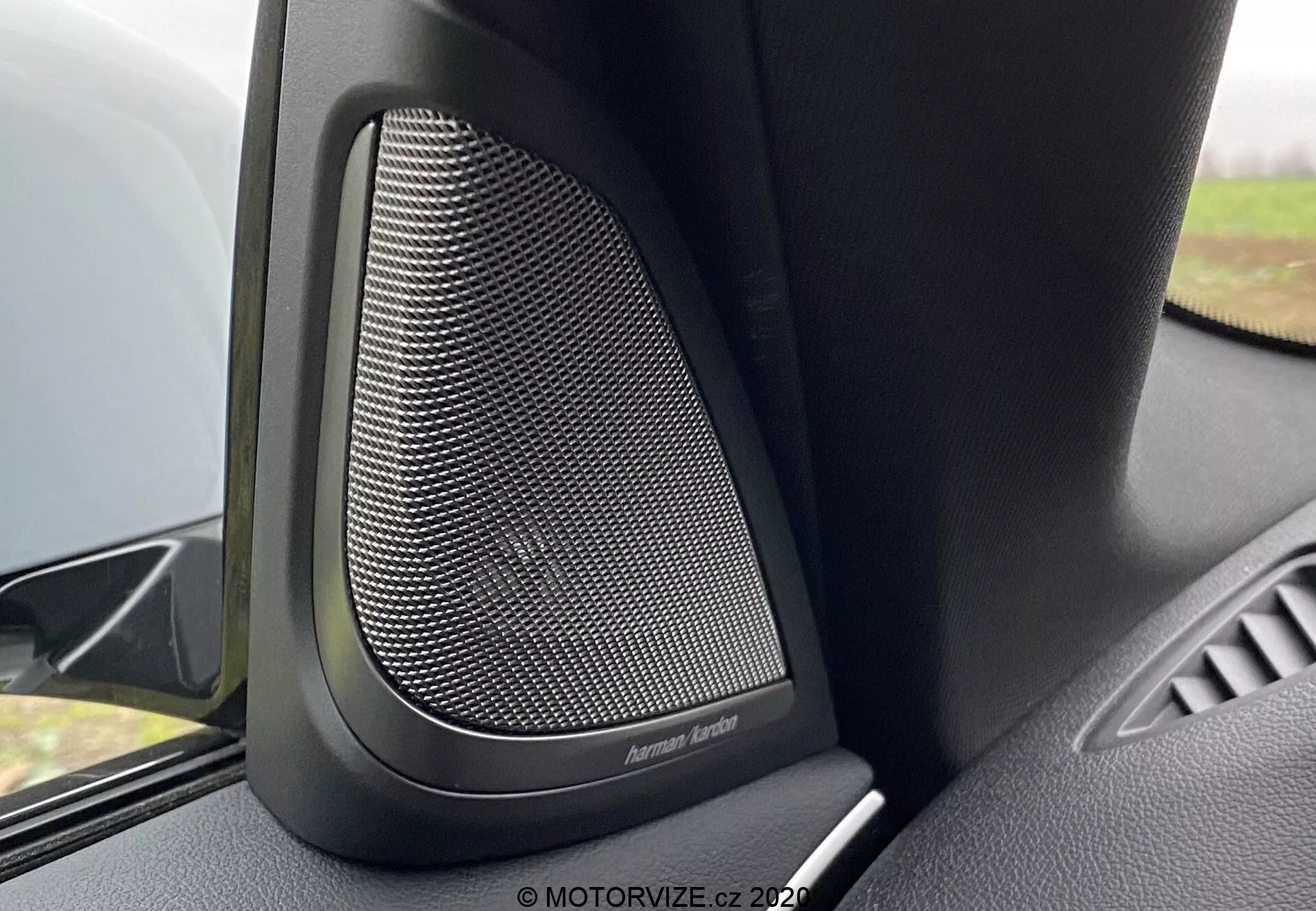 Närbild av interiören i BMW 5er-Reihe 2020 (BMW 520i Touring Steptronic) med fokus på dörrhögtalaren av premiumkvalitet från Harman Kardon. Bilden fångar det finmaskiga mönstret på den metalliska högtalargallret, omgiven av svart plast eller mjuka material som är utformade för att dämpa vibrationer och ge överlägsen ljudkvalitet. Kameravinkeln visar den övre delen av dörrpanelen, med högtalaren tydligt i fokus och en liten del av dörrens armstöd synlig. En mjuk bakgrundsoskärpa framhäver denna högkvalitativa ljudfunktion i detta lyxiga fordon.