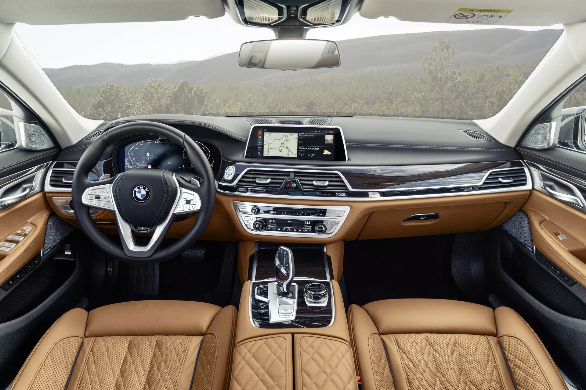 Bilden visar interiören av en BMW 7-serie 2019 (G11/G12 facelift) från ett bakåtsätesperspektiv, vilket framhäver den detaljerade och lyxiga designen på framsätet. Den moderna ratten med multifunktionsknappar och en framträdande BMW-symbol kompletteras av en elegant instrumentpanel med en central pekskärmsinfotainmentdisplay. Mittkonsolen innehåller en automatisk växelspak, pekplatta och olika dynamiska kontrollsystem, kompletterat av ljusbruna quiltade lädersäten som indikerar en premiumutrustningsnivå. Dörrpanelerna speglar denna lyx med ljusbrunt läder, träpaneler och ambient belysning, vilket antyder att avancerade teknik- och komfortfunktioner är integrerade i hela fordonet, inklusive ett soltak för extra naturligt ljus.
