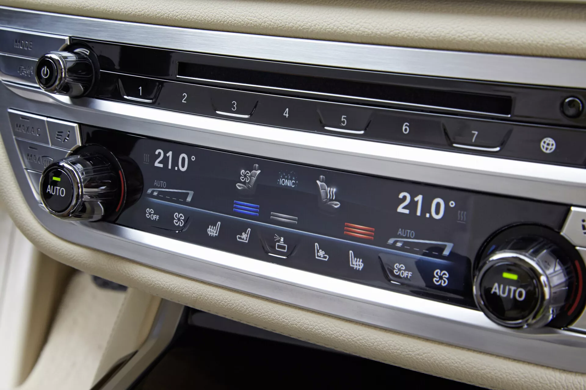 Närbild av BMW 7er-Reihe 2015 (G11-G12) klimatsystem på mittkonsolen mellan förar- och passagerarsätena. Fokus ligger på horisontell panel med två rattar och knappar för olika funktioner. Båda rattarna är inställda på 21,0°C och markerade som 'AUTO', vilket antyder tvåzons automatisk temperaturkontroll. Knapparna inkluderar bakrutedefrost, luftcirkulation, vindrutedefrost och ventilaktivering. Klimatsystemet är inramat av borstad metall mot en ljusinredning, med en möjlig display eller beröringskänslig remsa ovanför panelen.