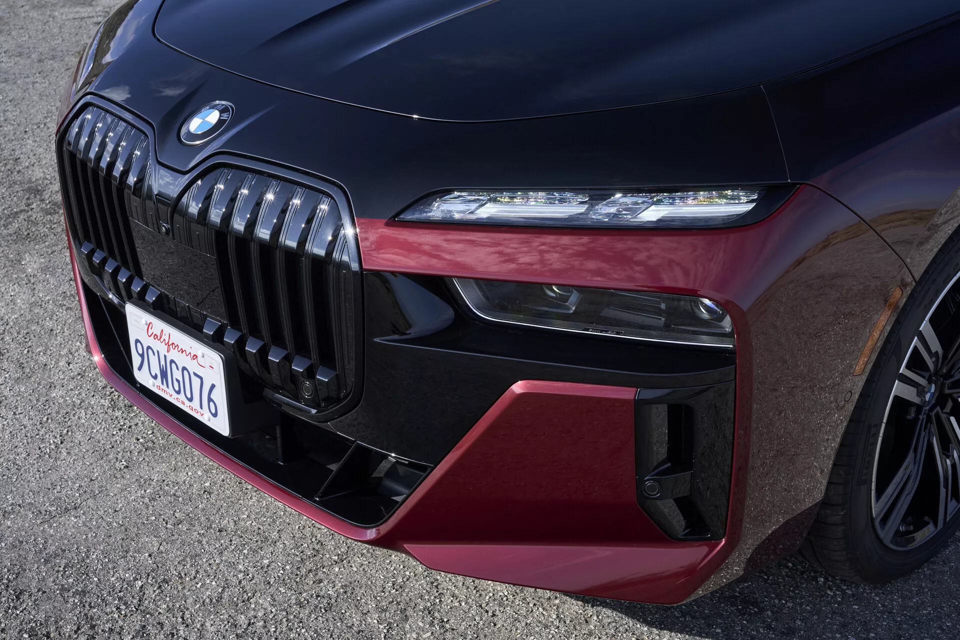 Närbild av den främre delen av 2023 BMW 7-serie (G70) som framhäver den kromade njurgrillen, eleganta LED-strålkastaren och den dynamiska frontstötdämparen med glansiga svarta och burgundy röda inslag, vilket understryker lyx och sportighet. Den låga vinkeln avslöjar en flerekrad lättmetallfälg i hörnet, som visar fordonets högteknologiska och prestandaorienterade egenskaper.